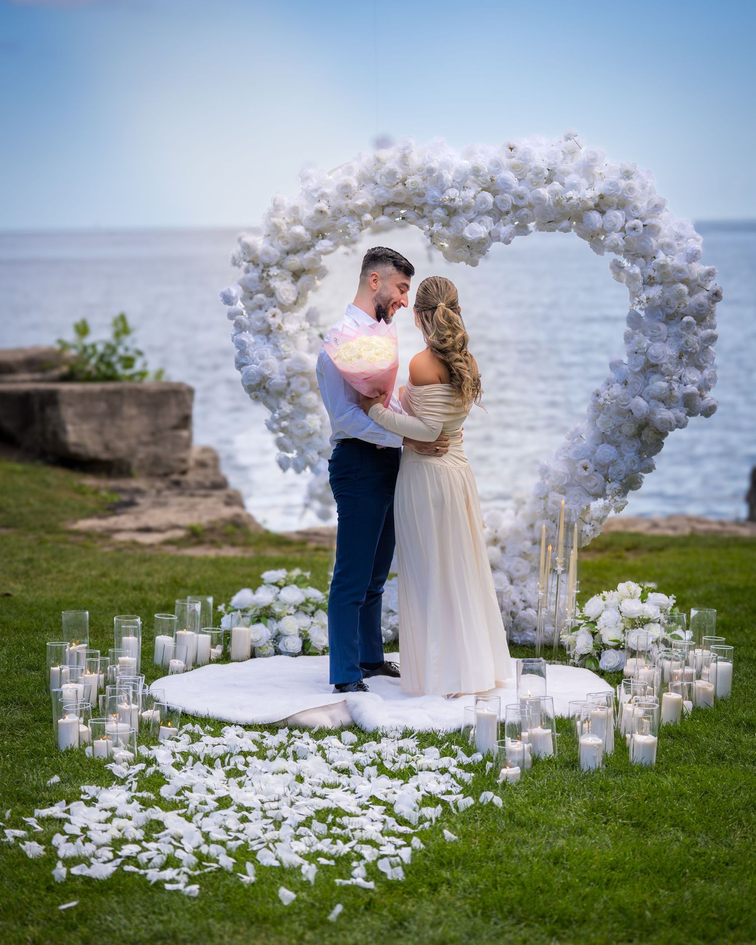 Seaside wedding ceremony site decorated with white floral arch and glowing candles in glass holders.
