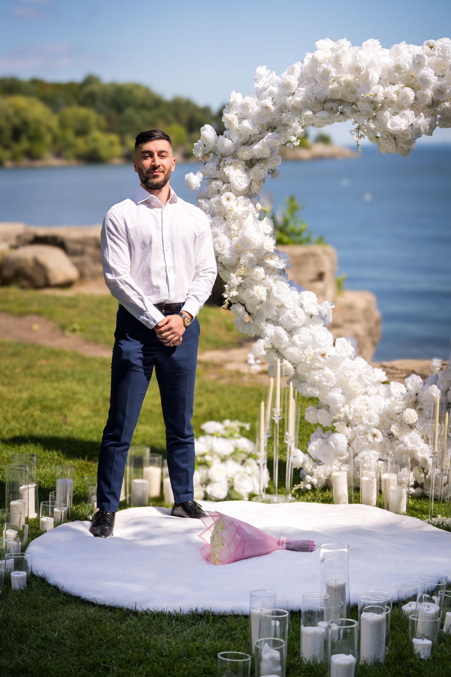 Wedding setup with white balloon arch and glass candle holders on grass by the water.