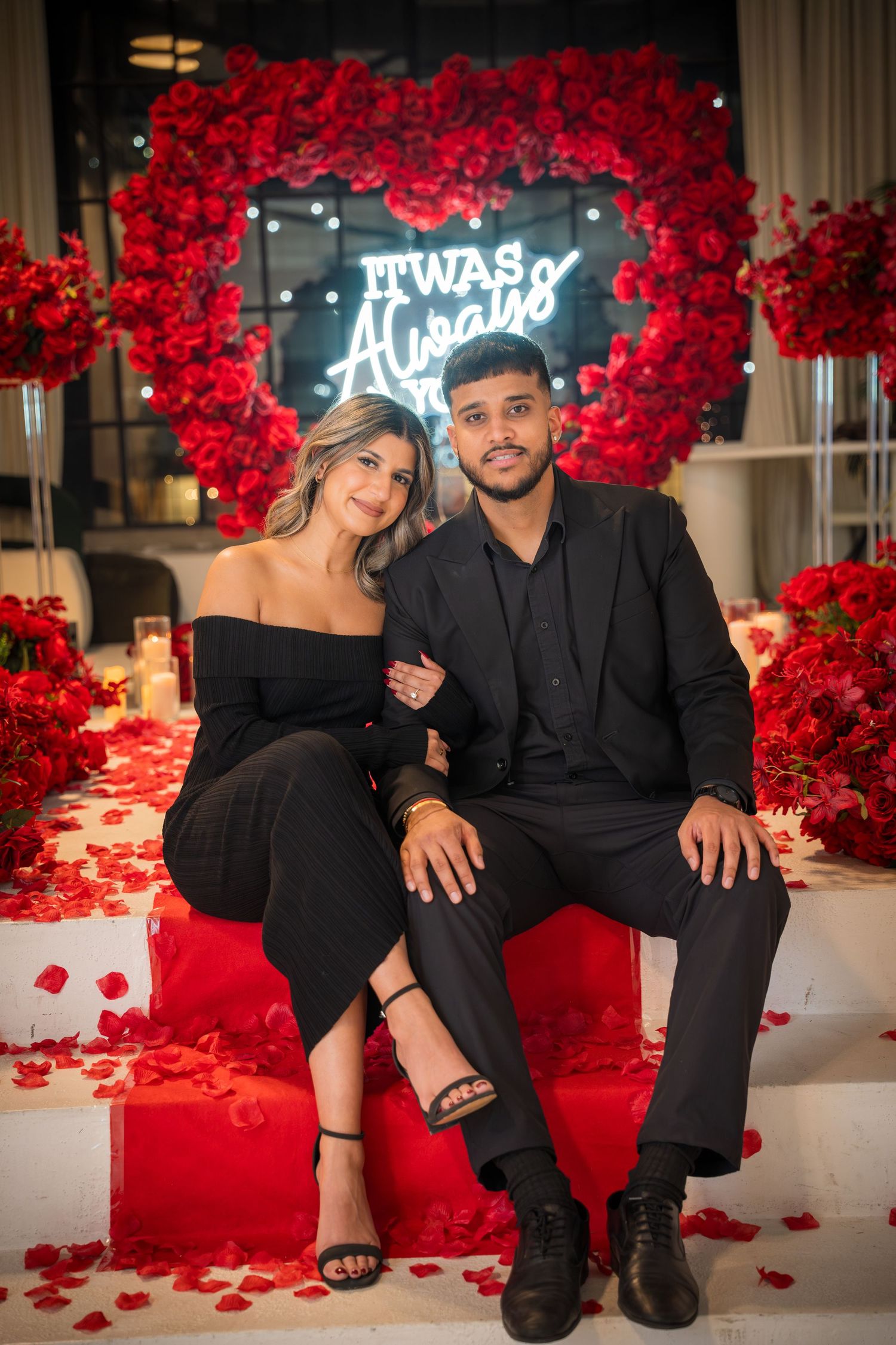 Romantic photoshoot featuring couple in black attire against wall of red roses and scattered petals.