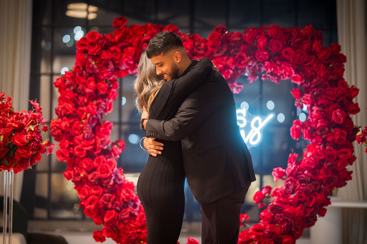 Couple celebrating their engagement in front of dramatic red rose backdrop.