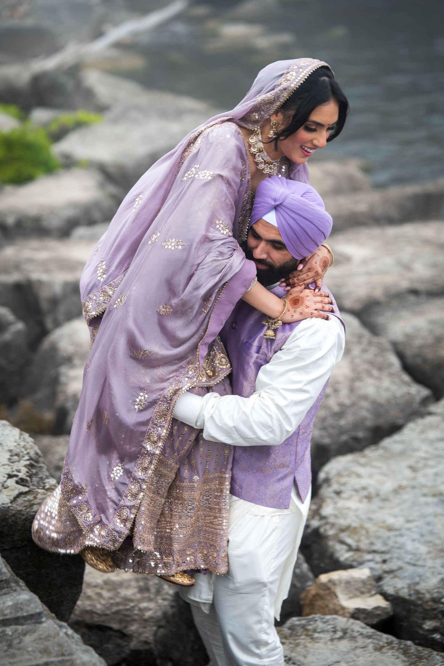 Couple in lavender and white outfits share intimate moments on rocky waterfront.