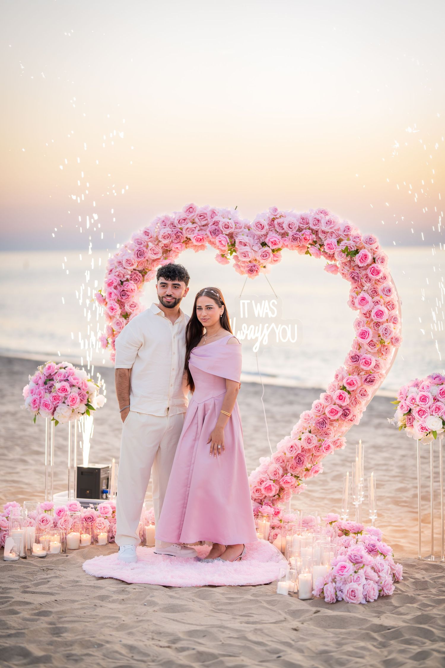 A couple stands under a pink rose heart-shaped arch on a beach during golden hour.