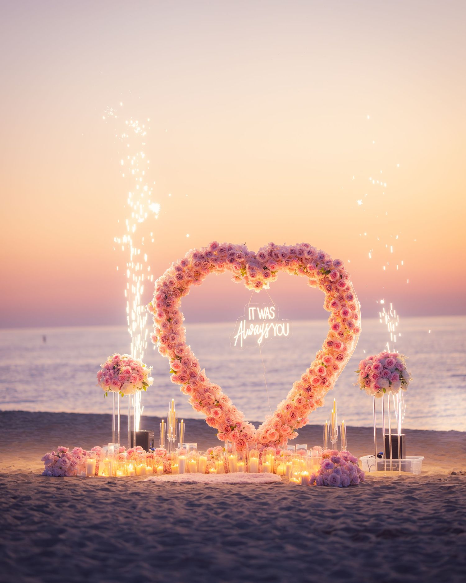 Romantic beach proposal setup with pink heart arch, sparklers and ocean view at dusk.