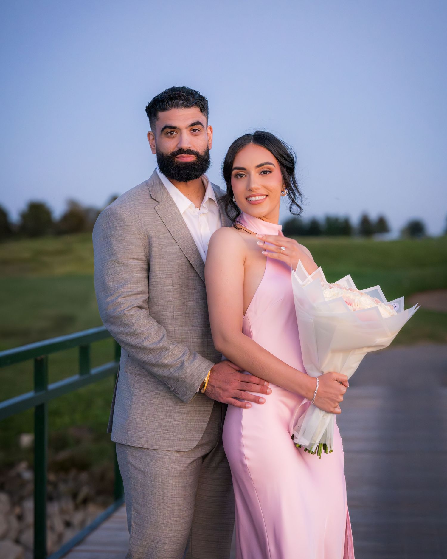 A couple in pink dress and gray suit pose with a white bouquet on an outdoor boardwalk.