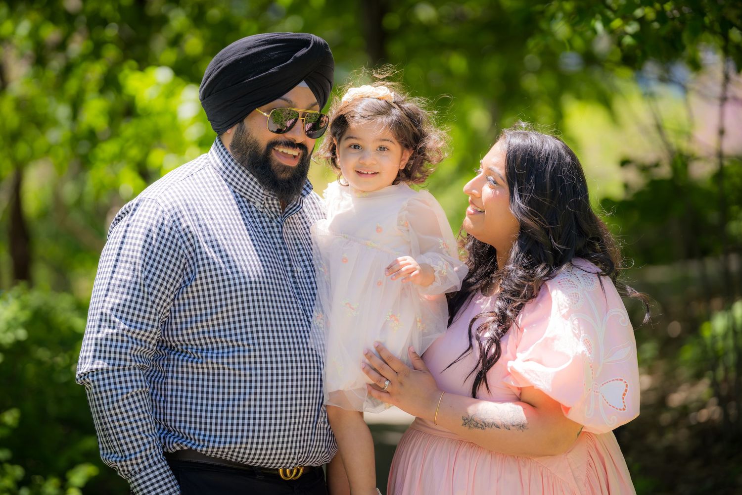 Sikh family of three with father in turban holding toddler in white dress, mother in pink gown.