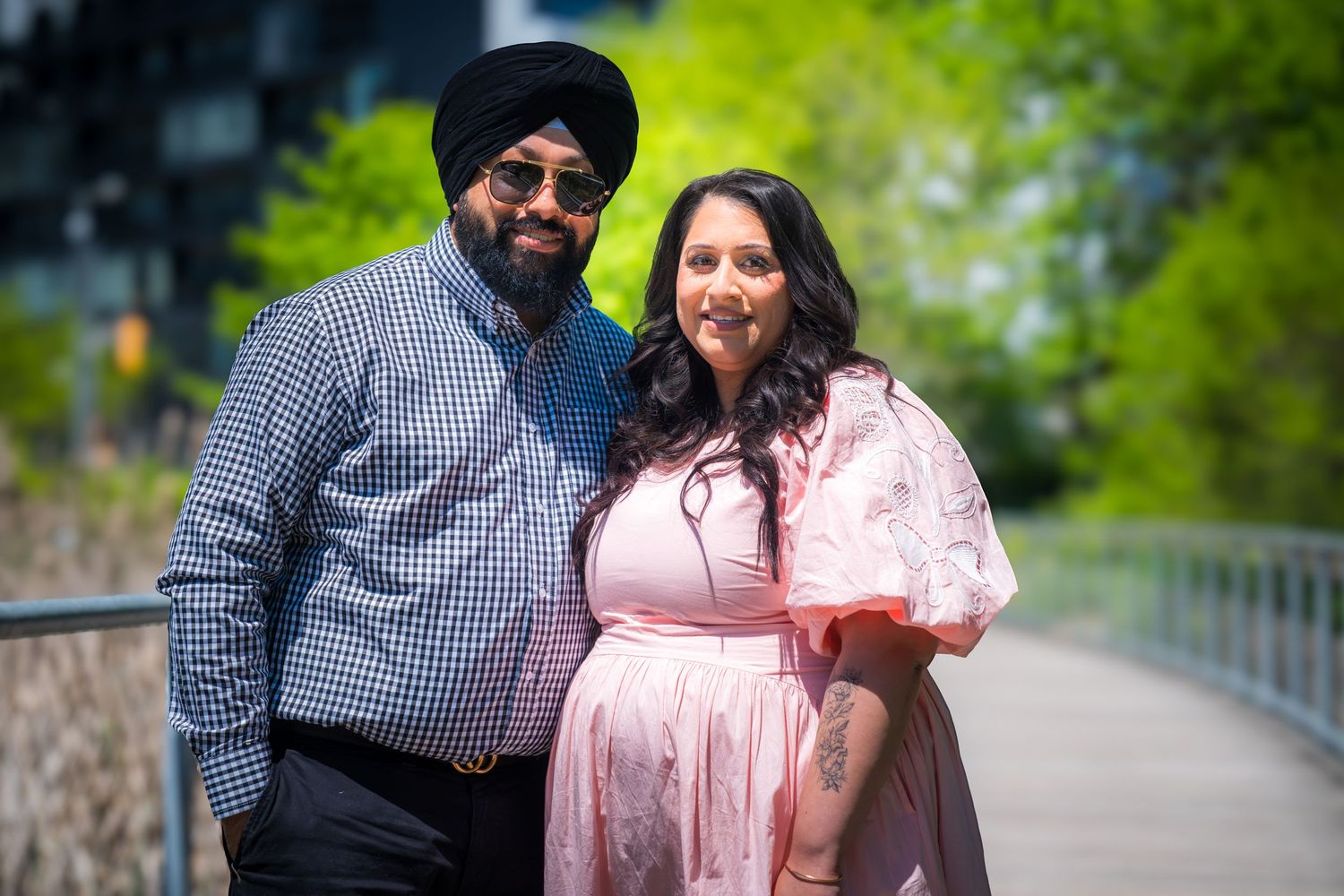 Sikh man in turban and sunglasses poses with woman in pink dress on a sunny outdoor walkway.