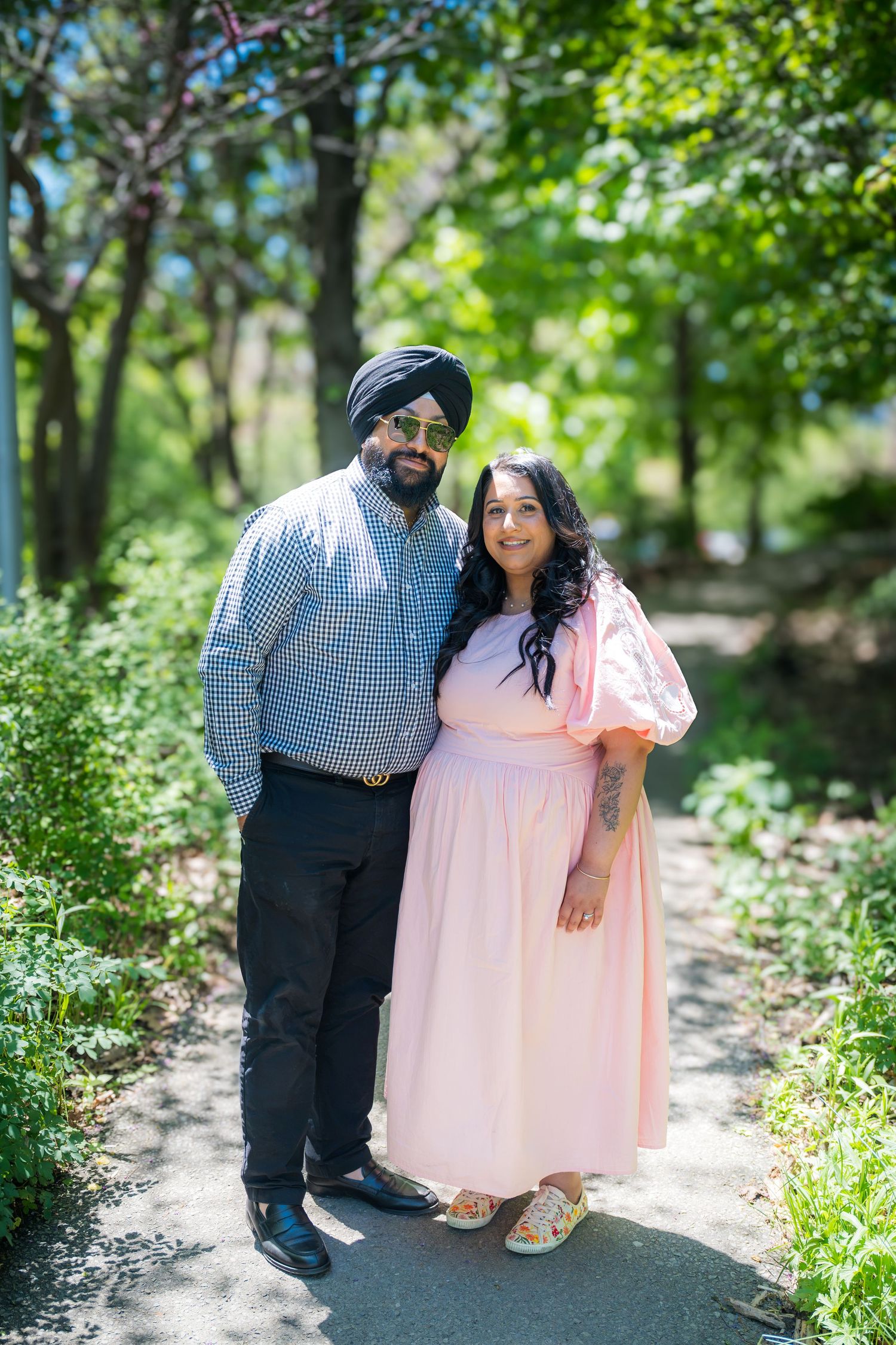 Sikh couple smiling together on a sunny park path surrounded by lush greenery.