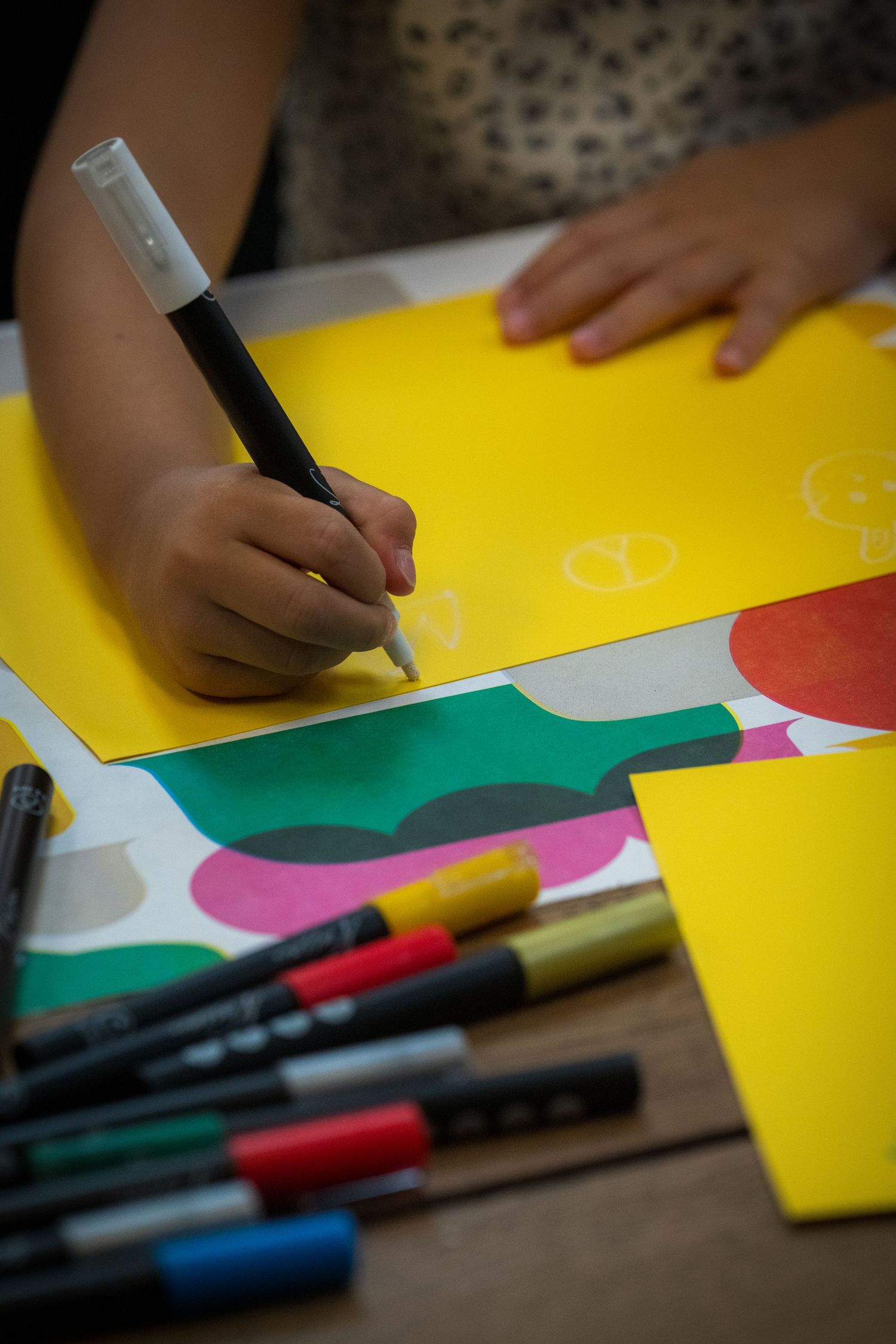 Child drawing with black marker on colorful yellow paper surrounded by multicolored markers on wooden table.