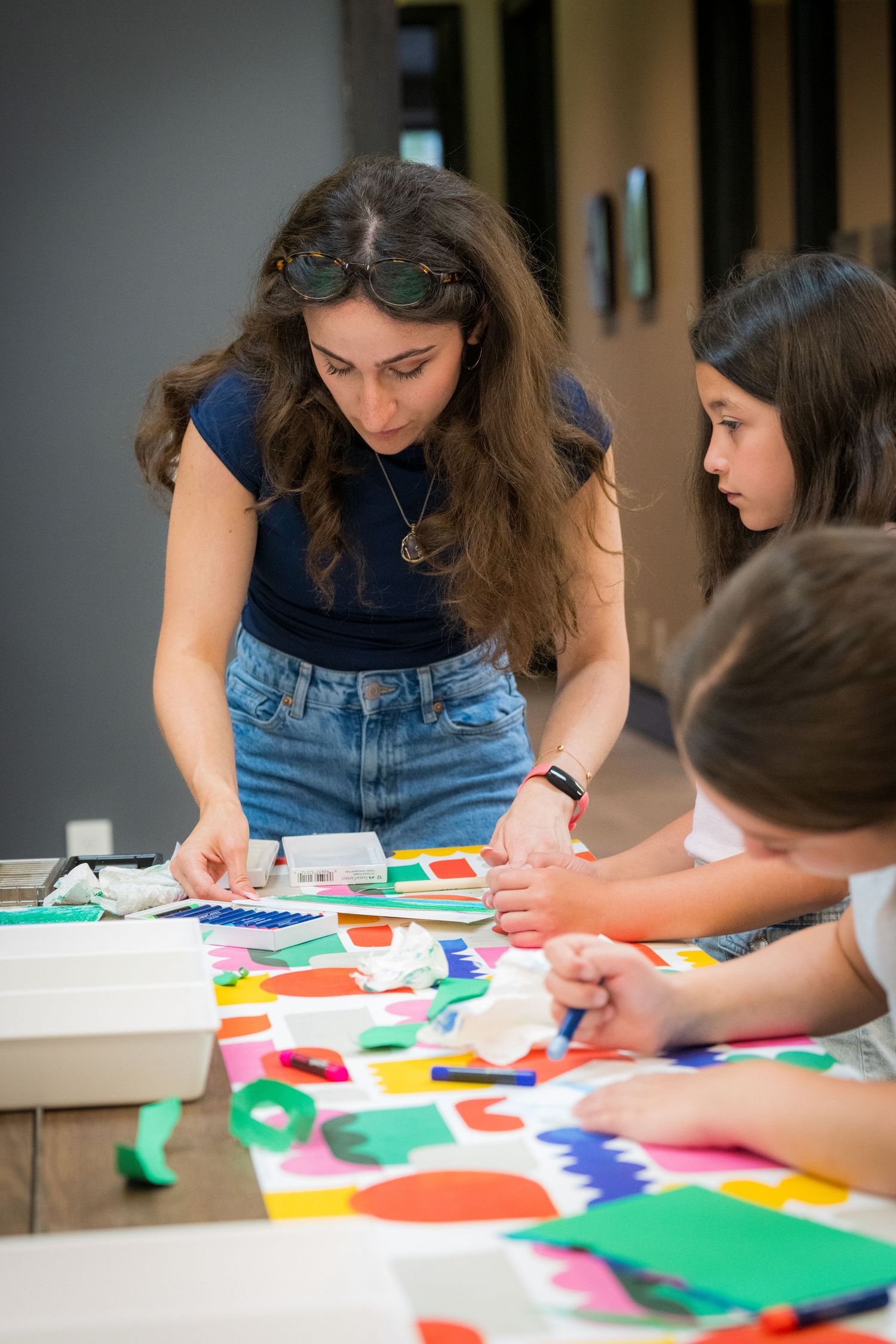 Female instructor guides two children working on colorful paper shape collage art project indoors.