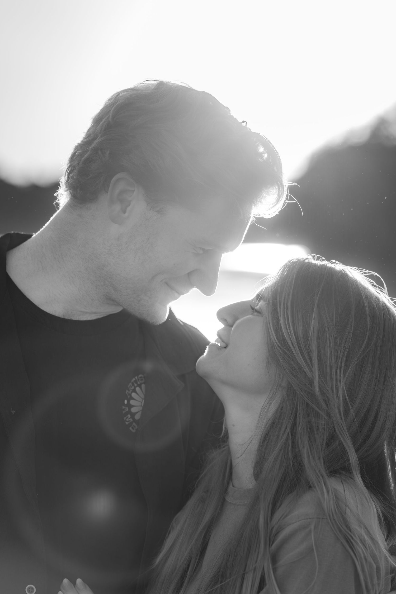 Black and white portrait of a couple leaning in for a kiss with bright sunlight behind them.
