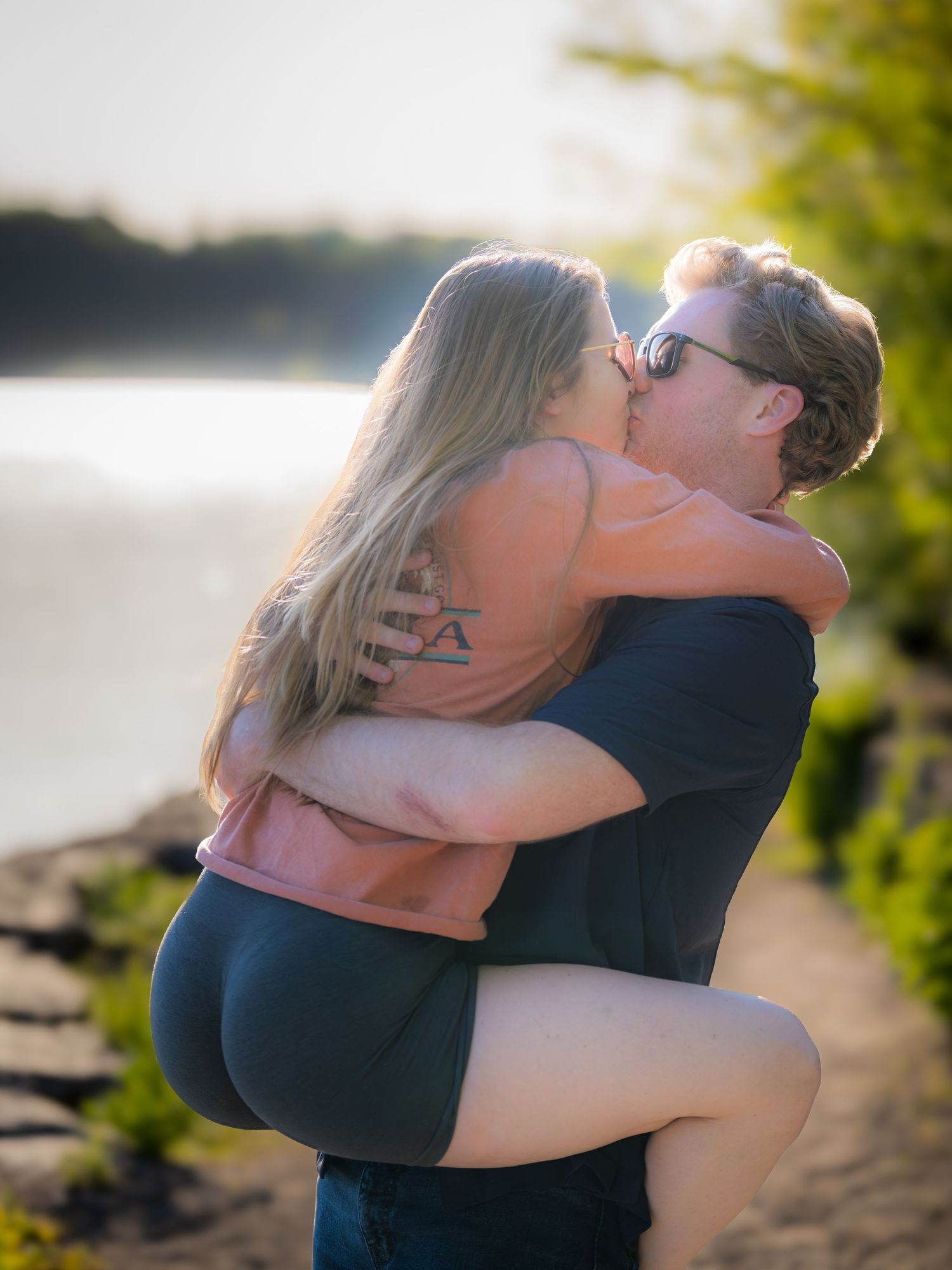 Couple sharing a passionate kiss outdoors near a lake at golden hour, man lifting woman in his arms.