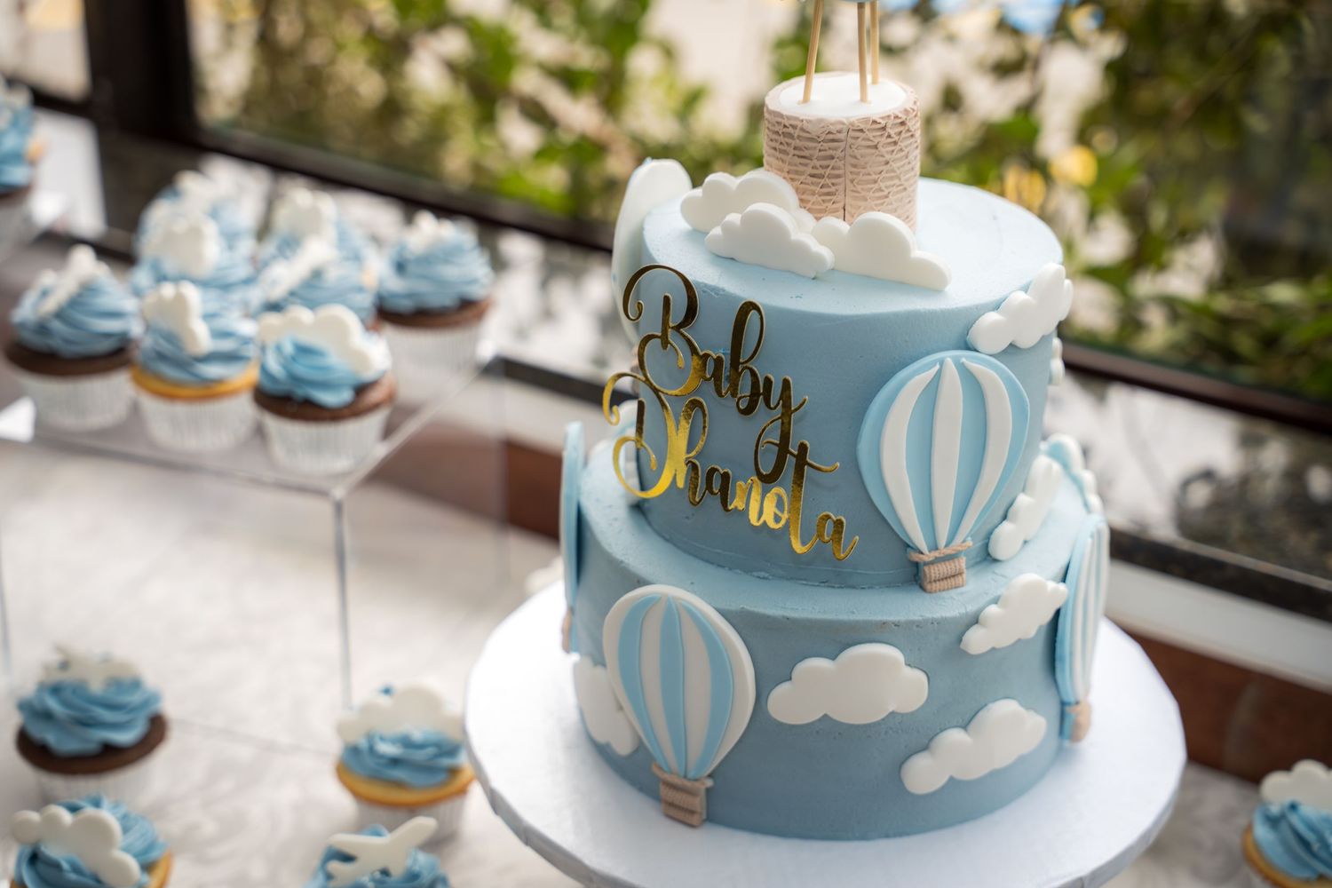 A baby blue and white hot air balloon themed dessert table with cake, cupcakes and decorative elements.