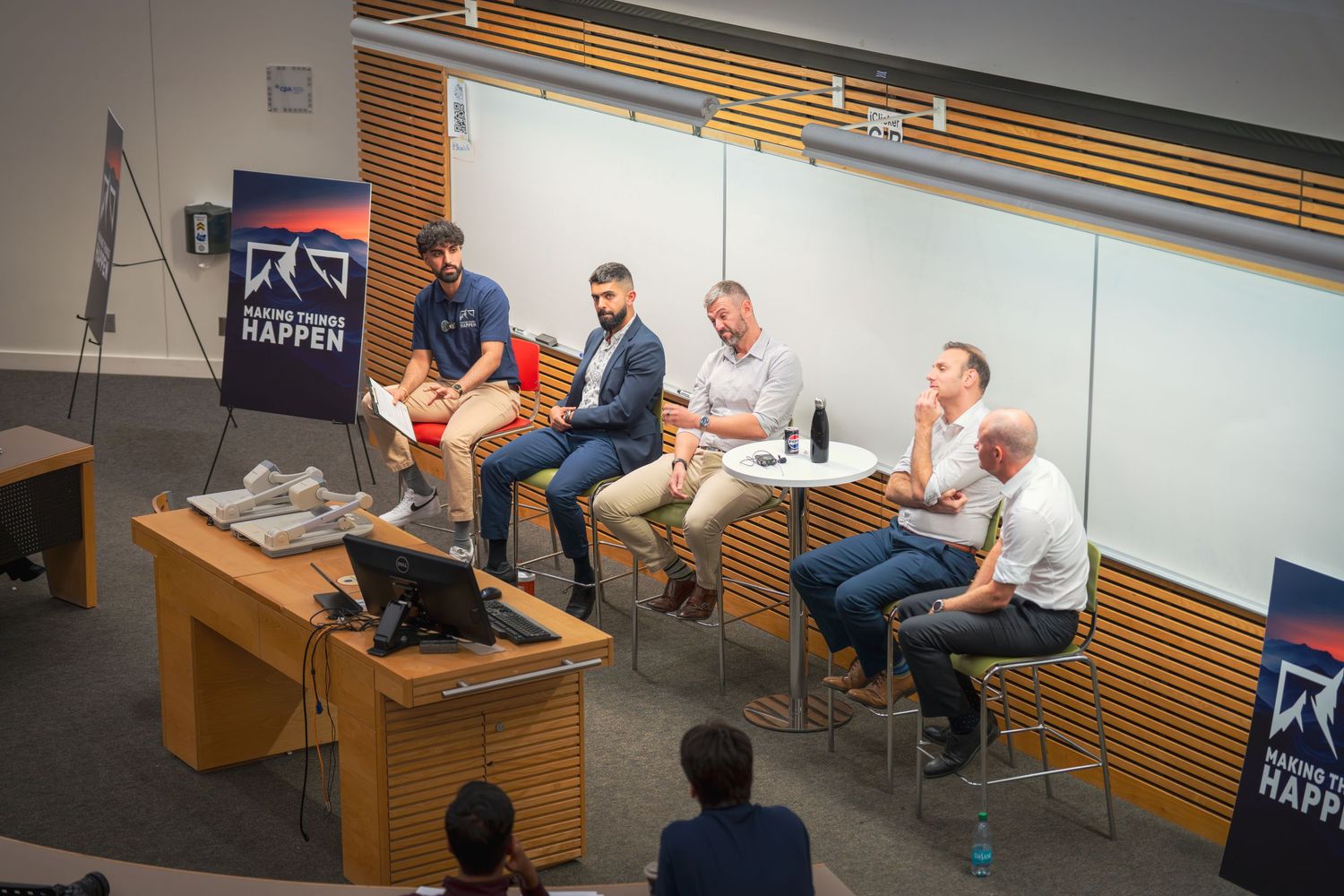 Panel discussion with five professionals seated in a modern conference room near a 'Make Things Happen' banner.