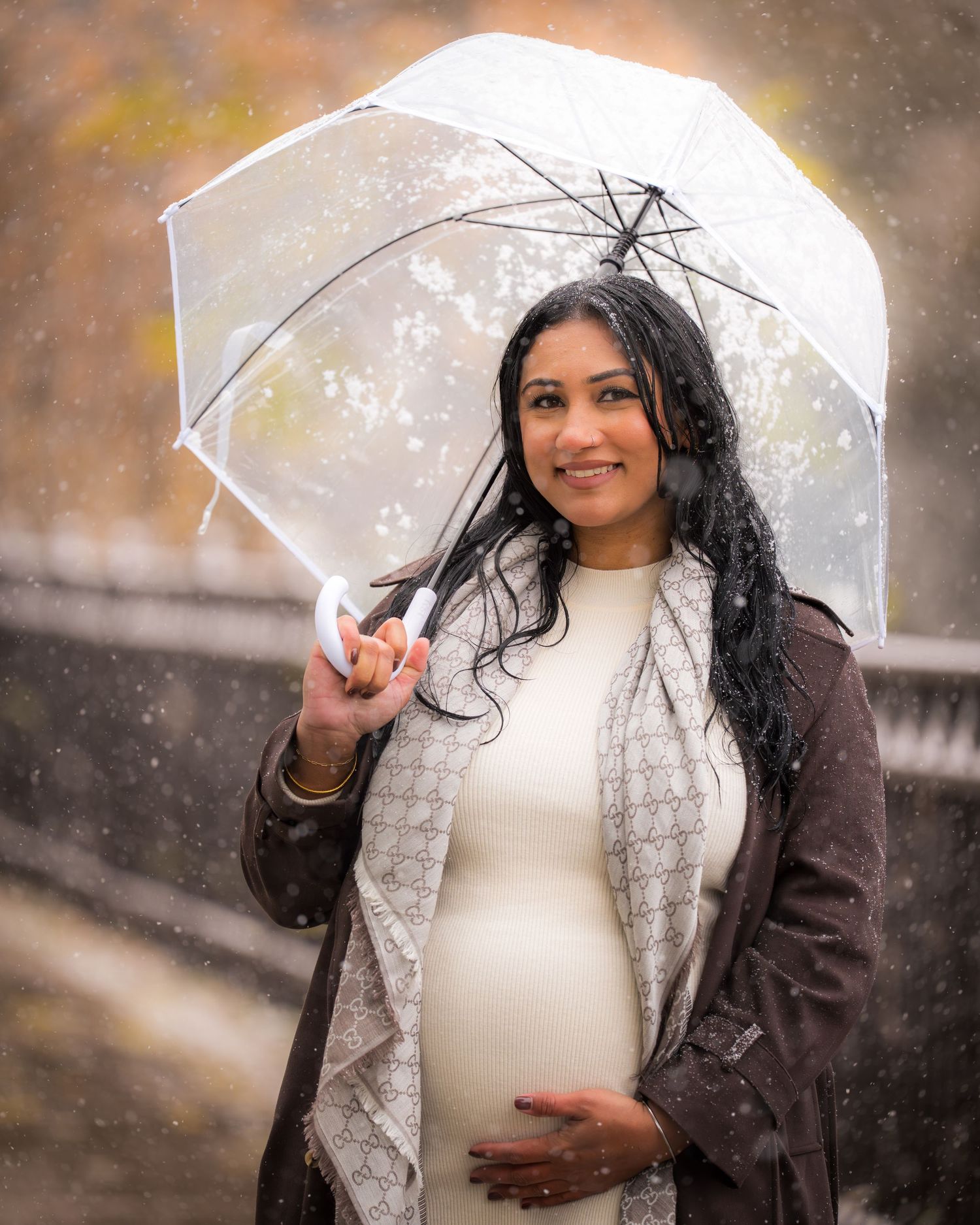 A person in a white sweater and brown coat holds a clear umbrella in falling snow.