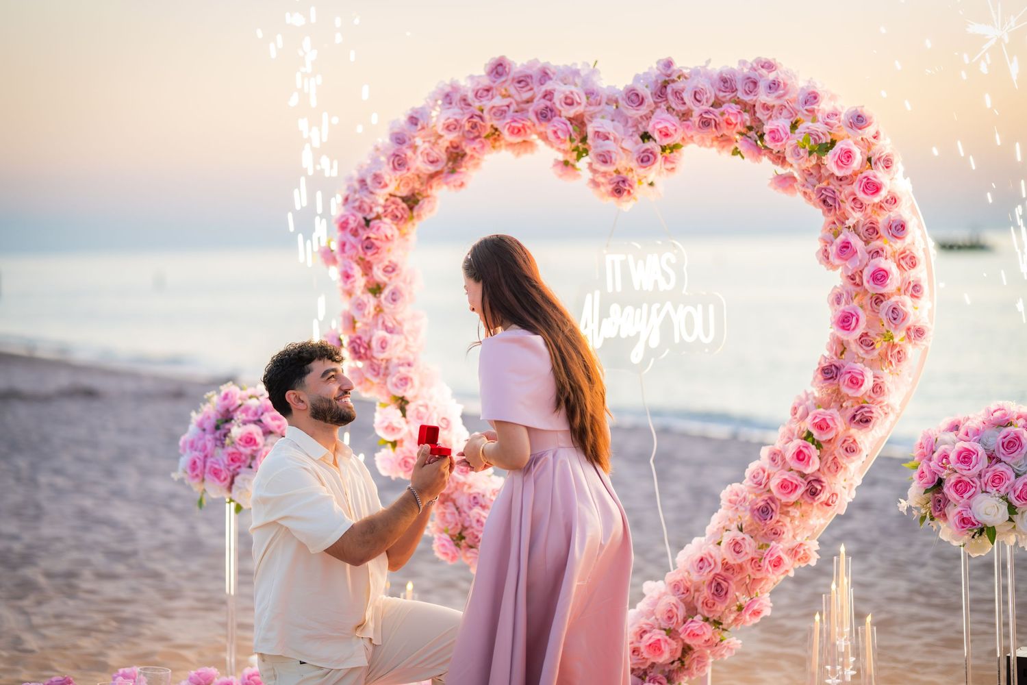 A romantic beach proposal moment captured under a pink rose heart arch at sunset.