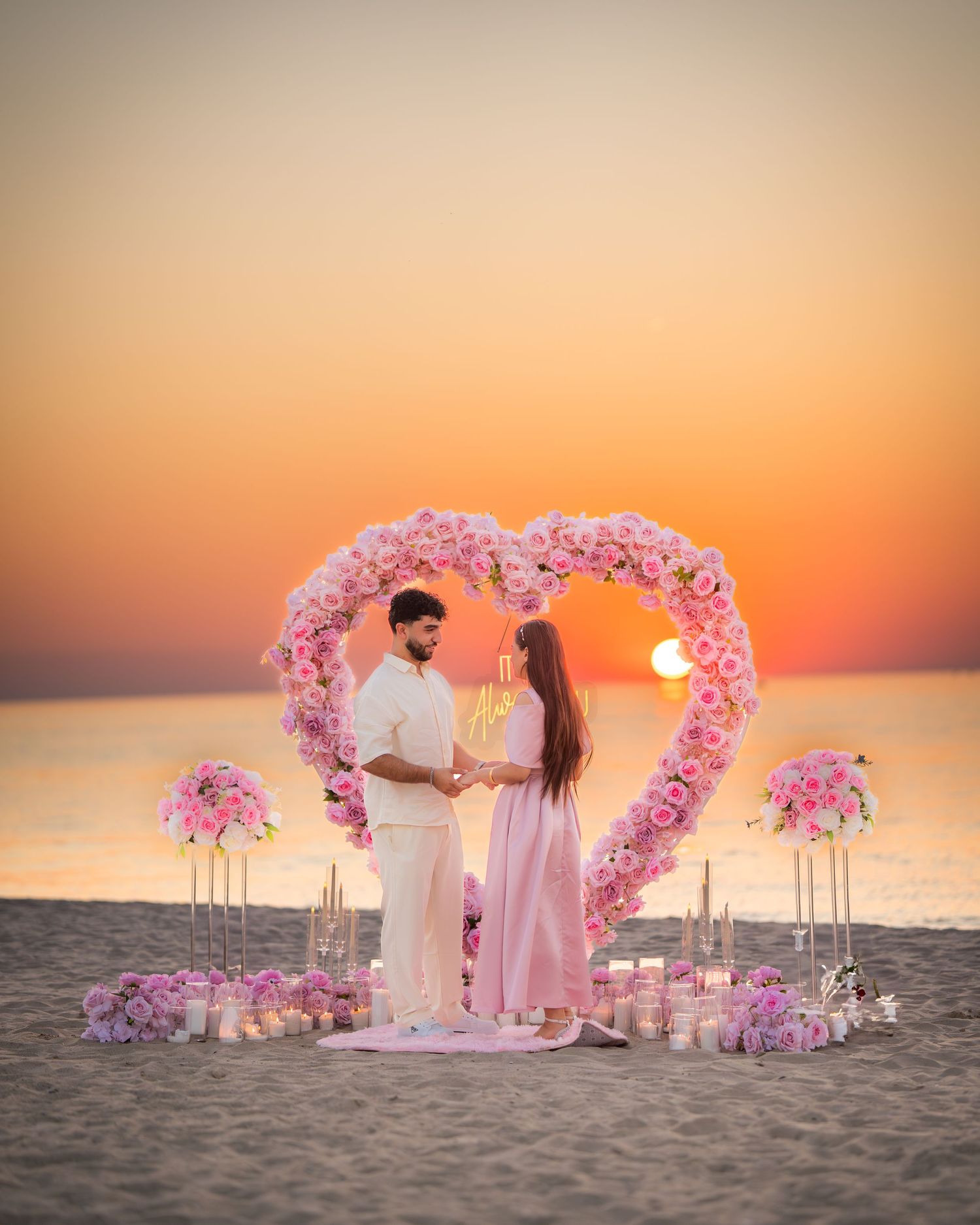 Pink floral heart arch setup on beach with romantic sunset and ocean view.