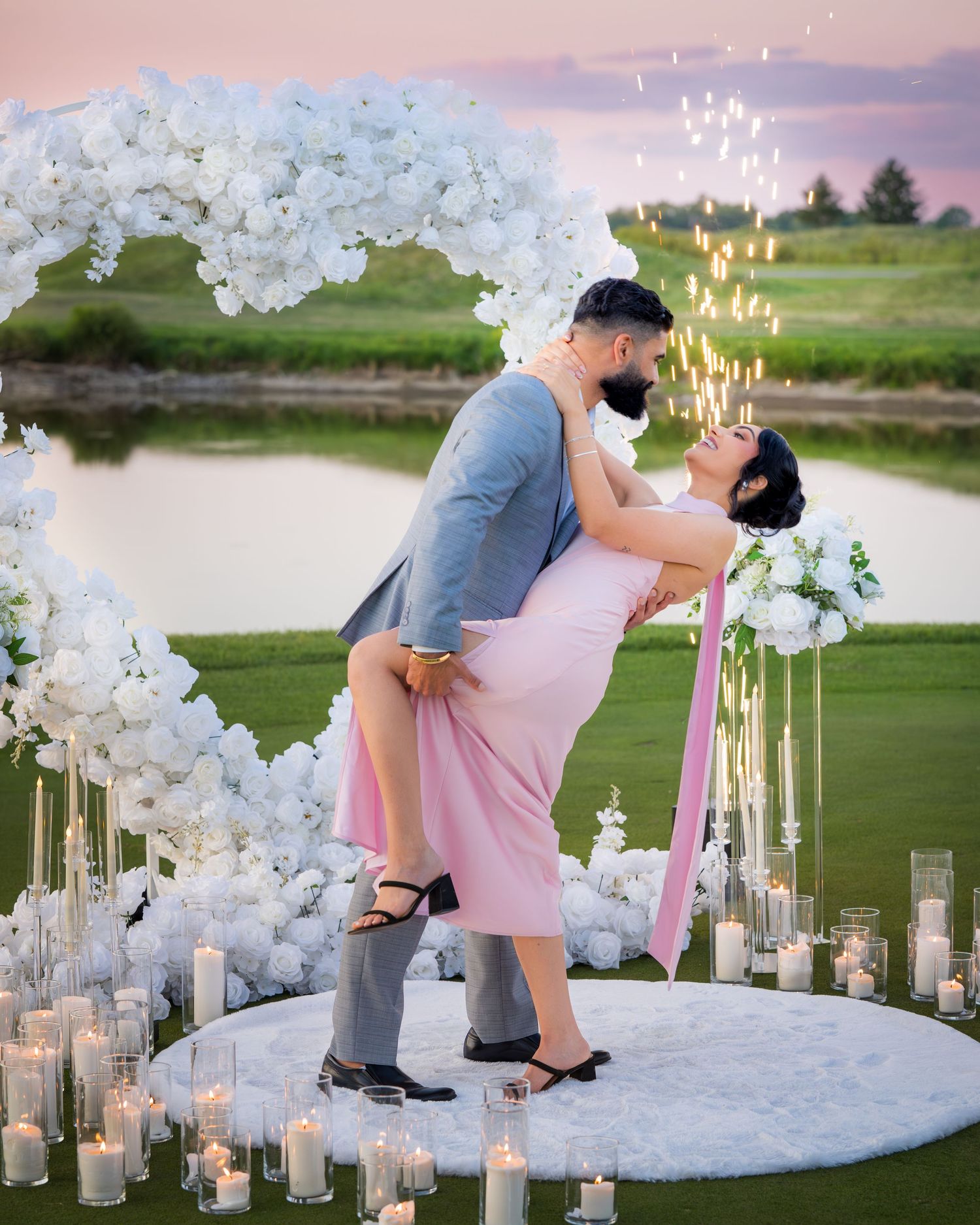 A couple performs a romantic dip pose surrounded by white flowers and lit candles outdoors.