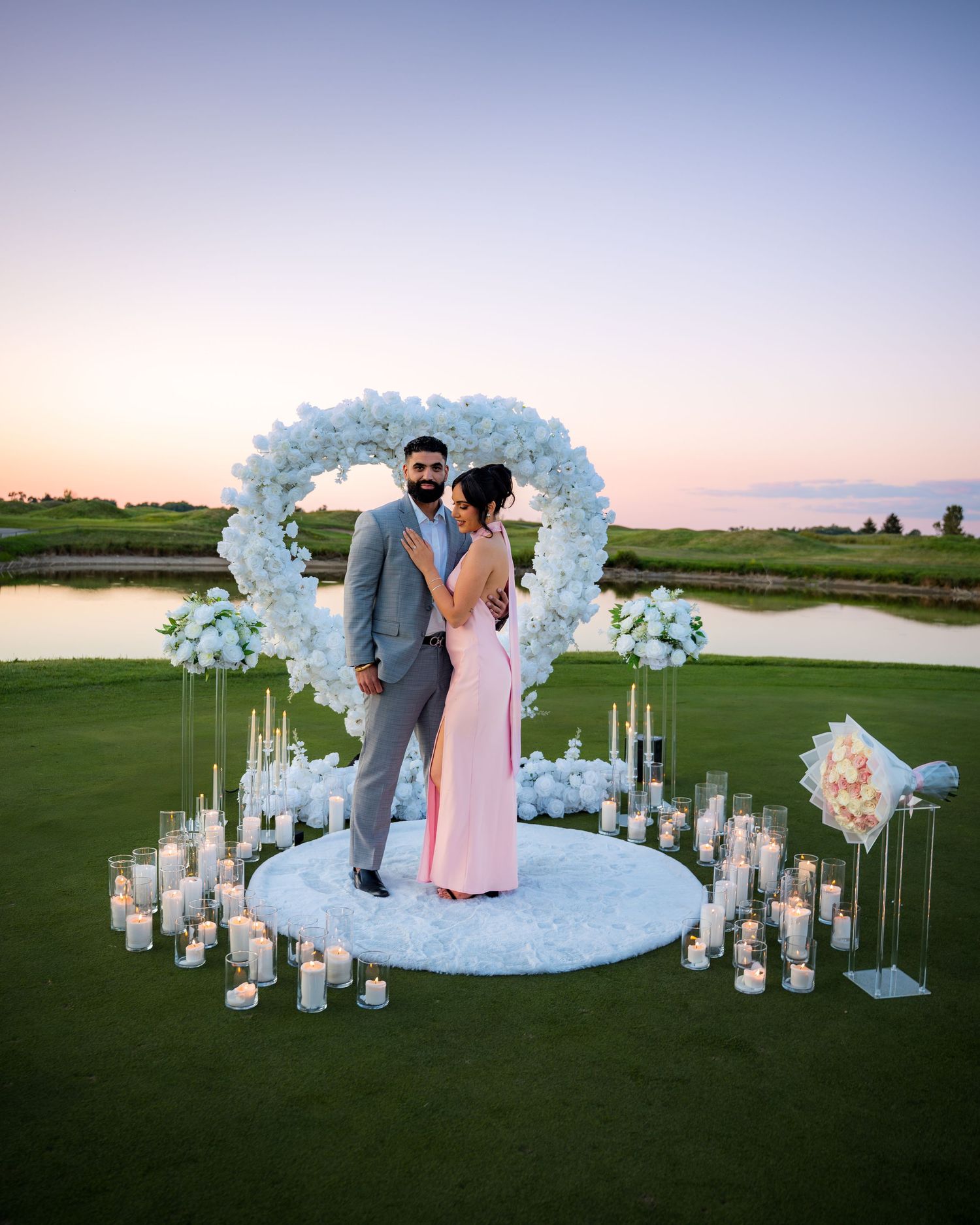 Romantic outdoor wedding setup with floral arch and lantern-lined aisle against sunset sky.