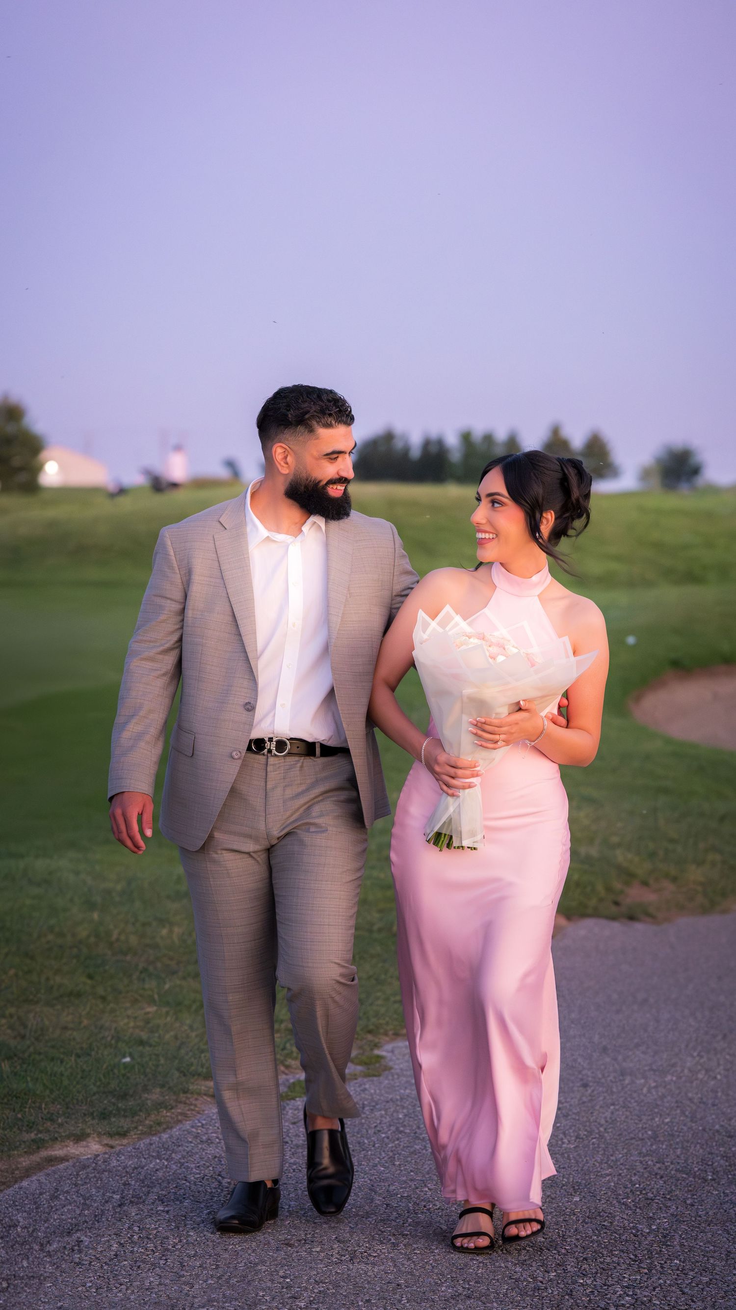 A romantic couple walks hand in hand on a path, wearing a grey suit and pink dress.