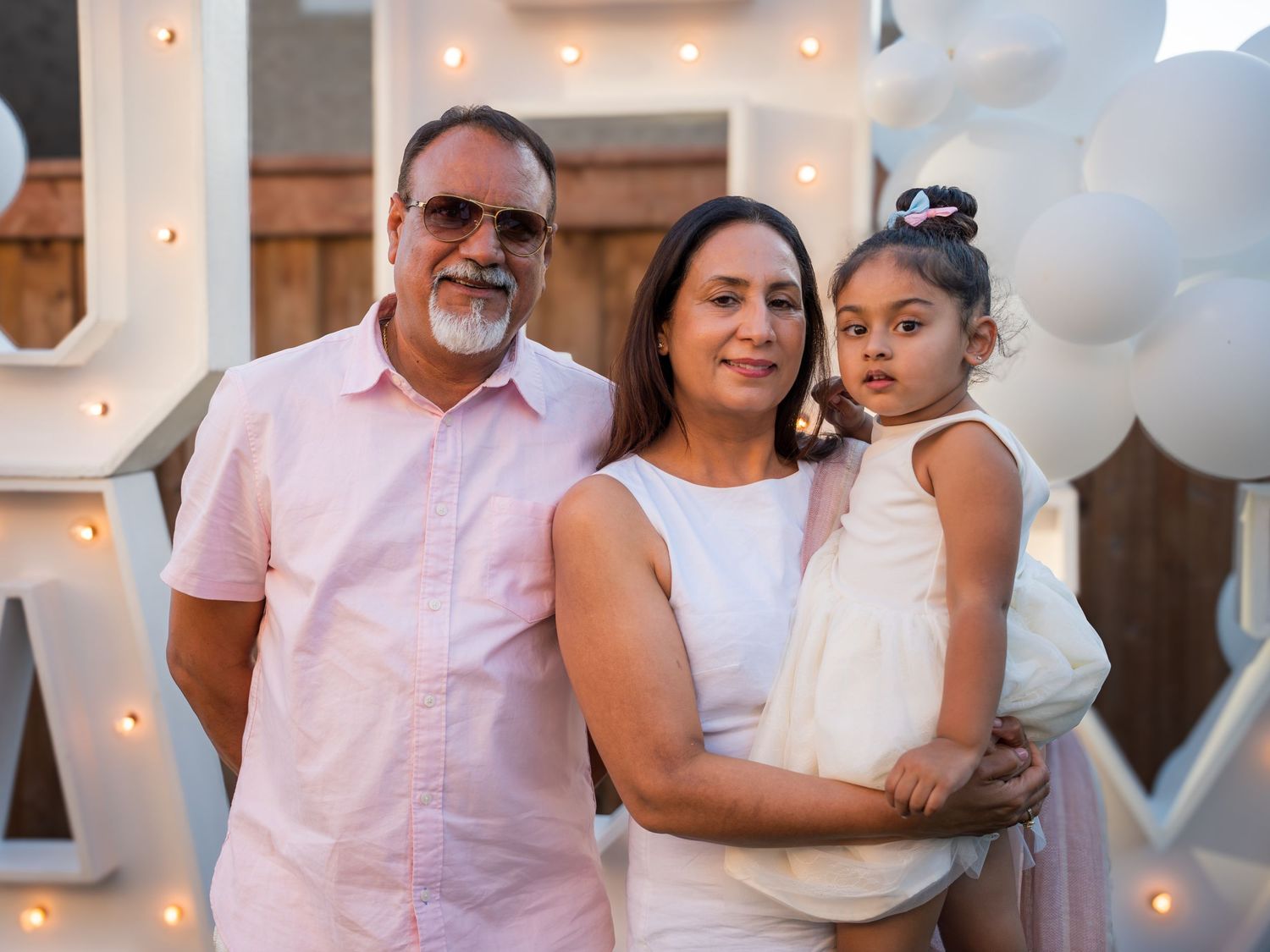 A family of three poses together at an outdoor party, with a man, woman, and young girl in white.