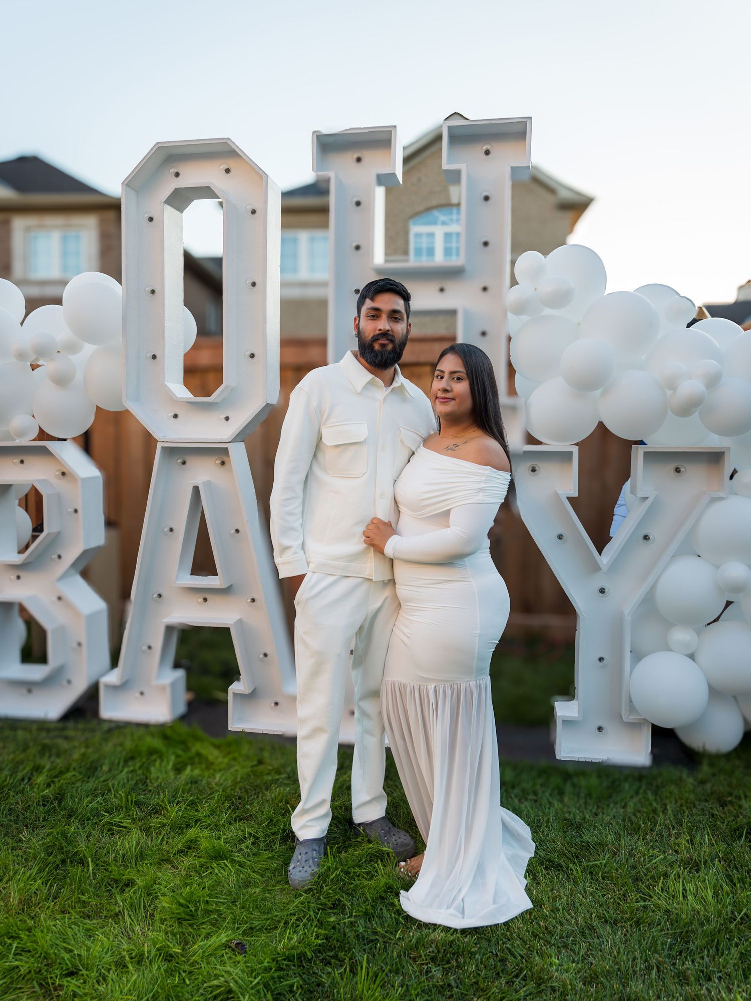 Expecting couple poses in front of illuminated OH BABY marquee letters with white balloon decorations.