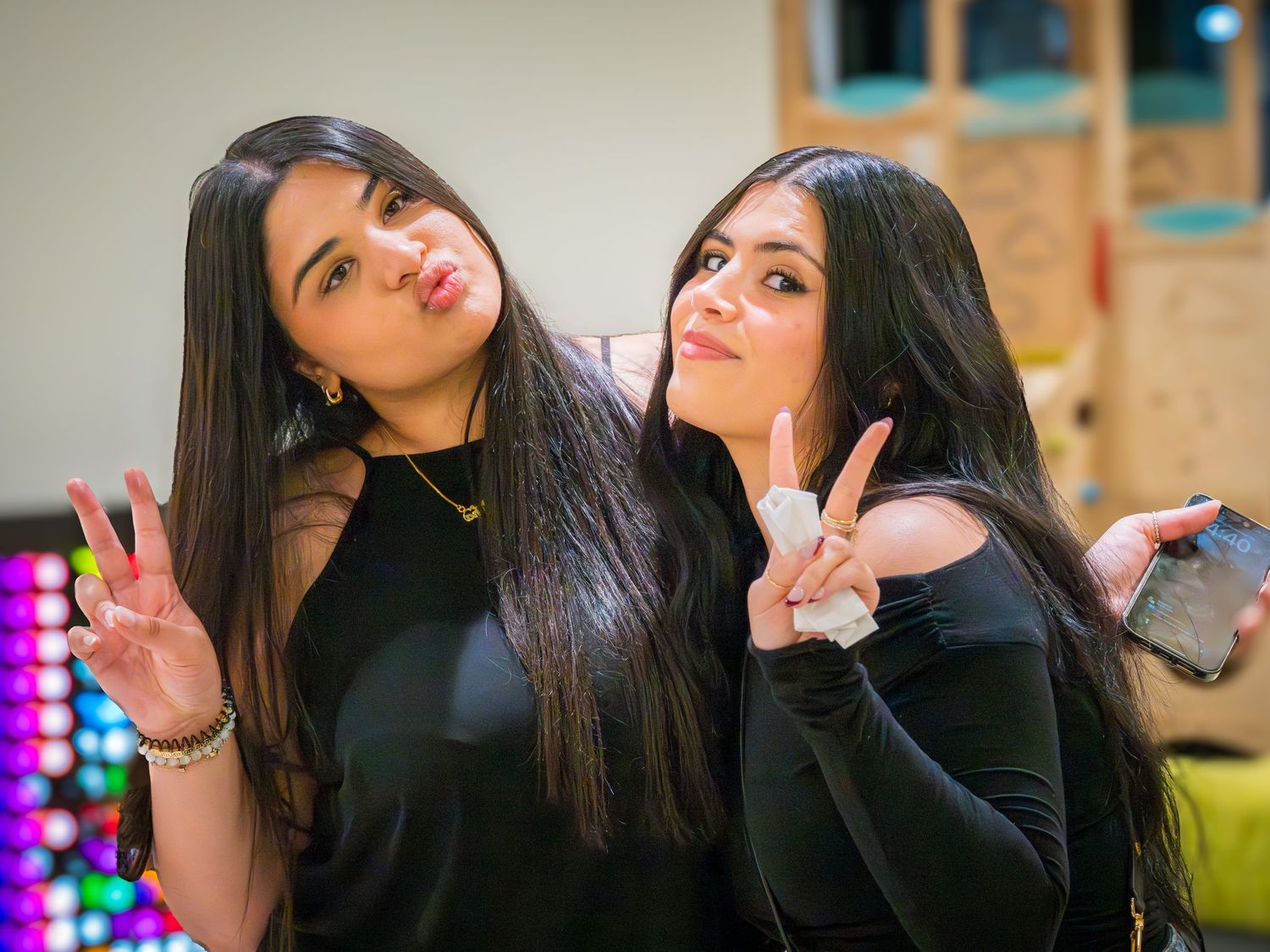 Two young women with long dark hair making peace signs and duck faces at a party.