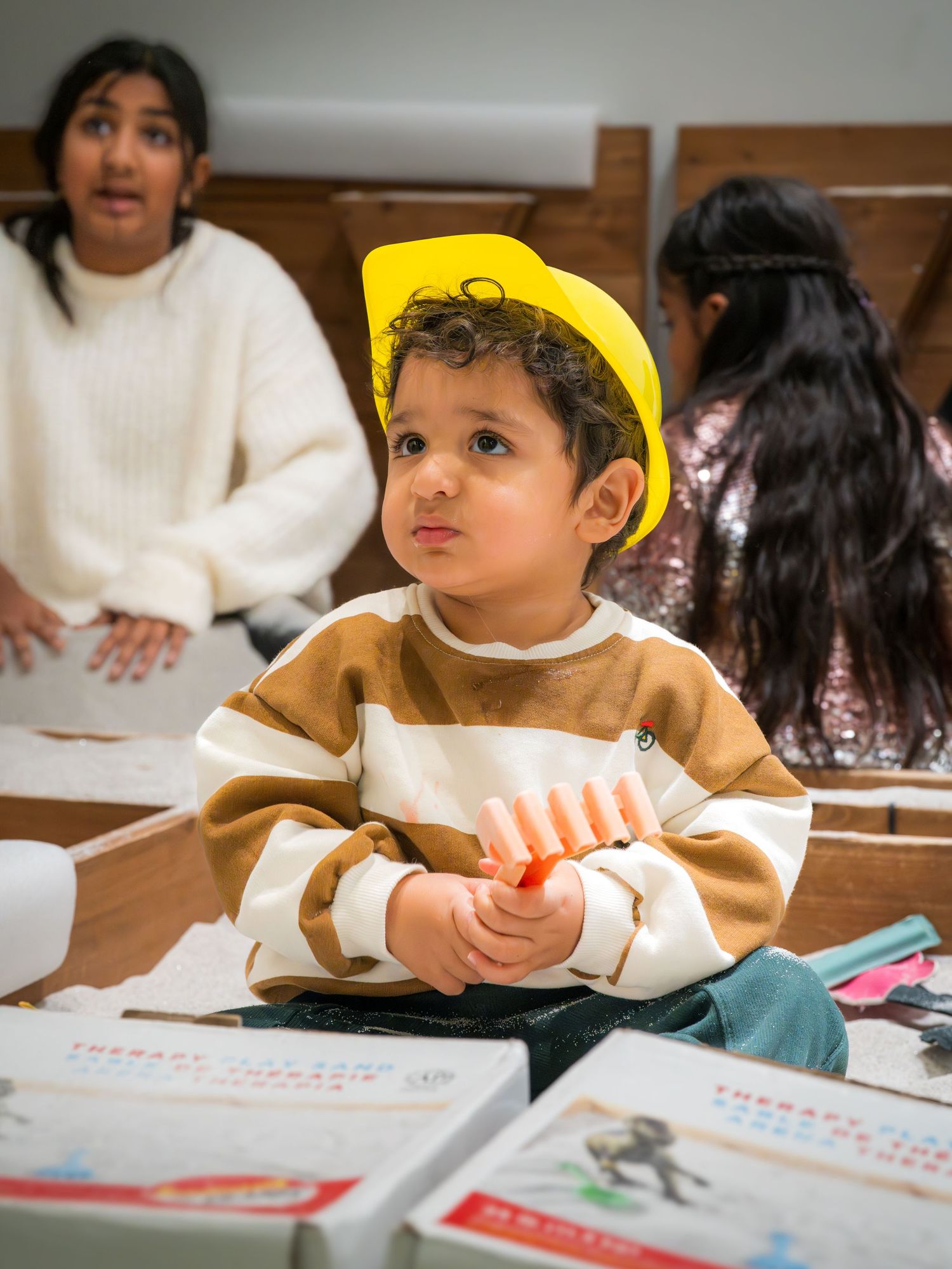 Cute toddler wearing a yellow paper hat sits at a table holding crayons during a craft activity.