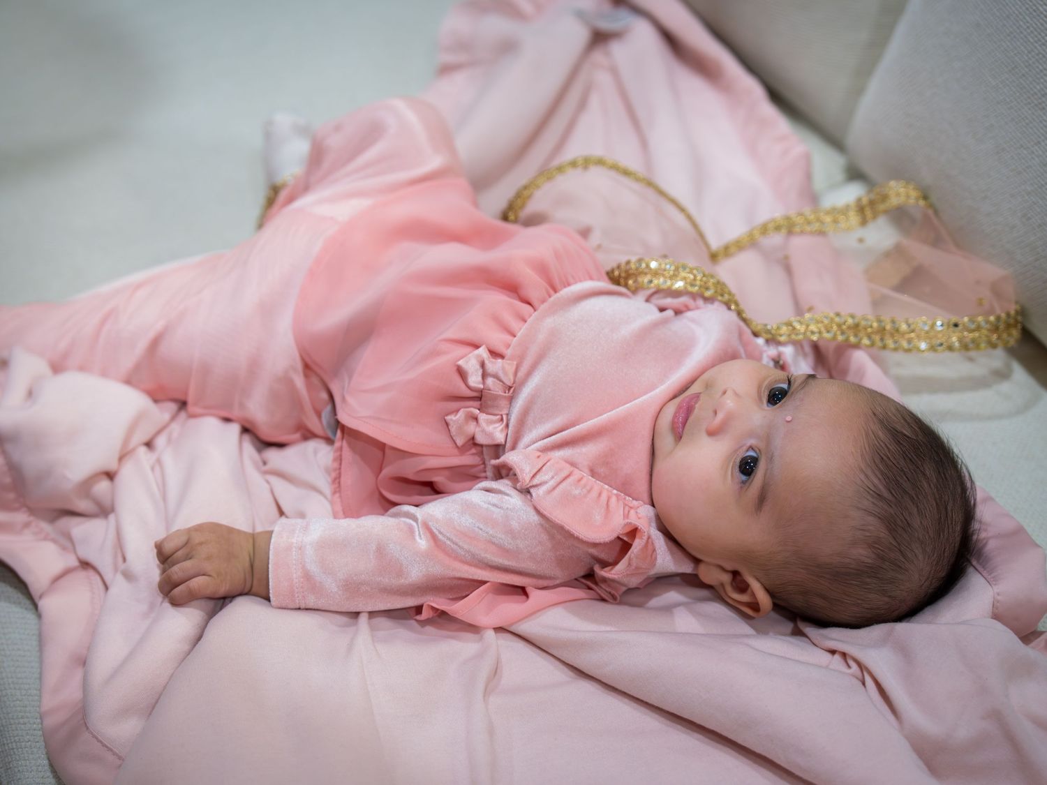 Newborn baby girl in pink ruffled outfit lying on pink blanket with gold ribbon decorations.
