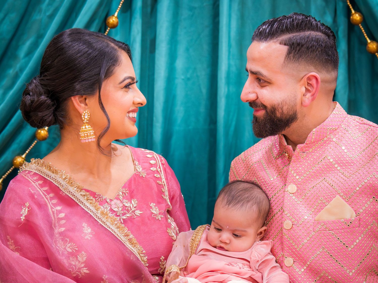 Indian couple in pink traditional outfits lovingly gazes at baby during cultural celebration event.