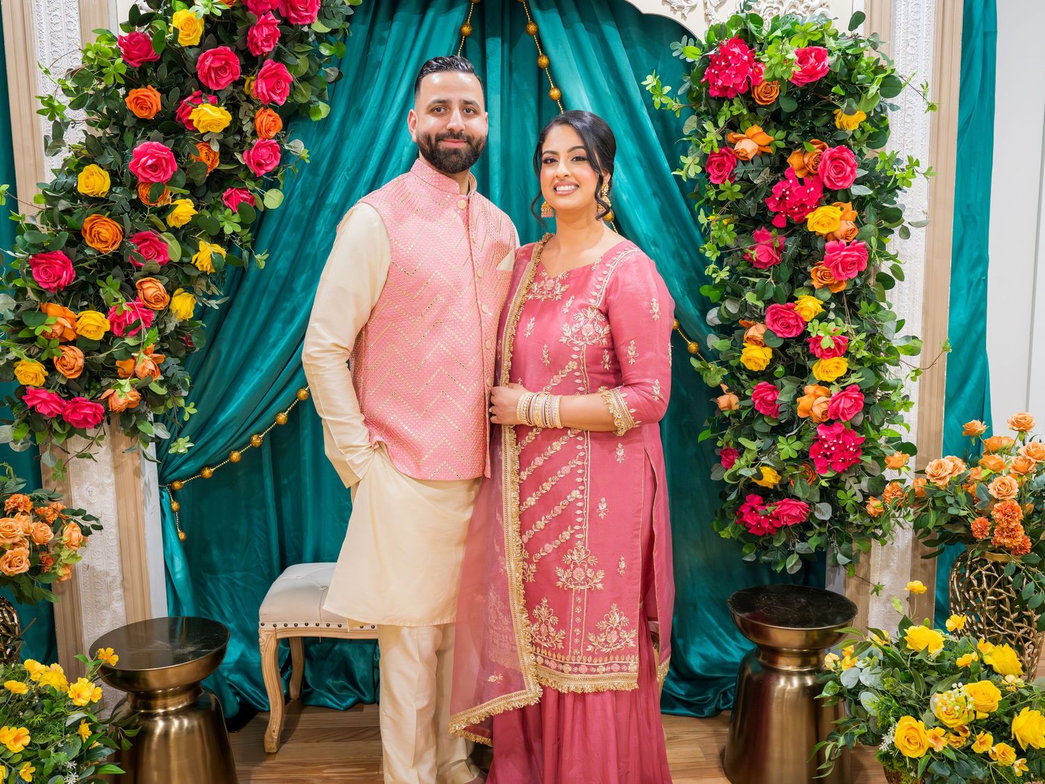 South Asian couple in pink traditional Indian outfits standing before colorful floral ceremony backdrop.