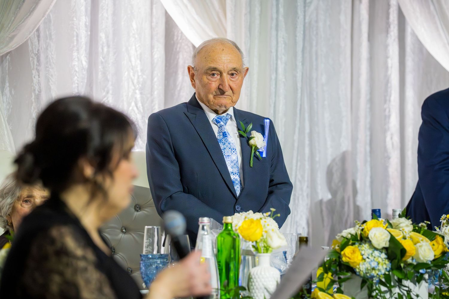 Elderly man in blue suit with boutonniere listening attentively to speeches at his anniversary celebration dinner.