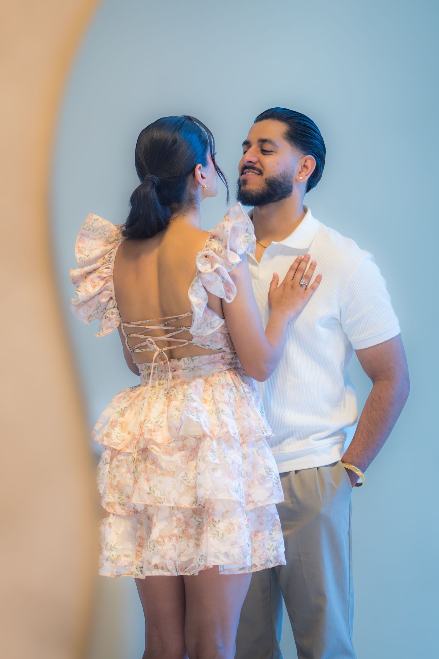 Couple in elegant proposal setting, woman in backless floral dress facing bearded man in white polo shirt.
