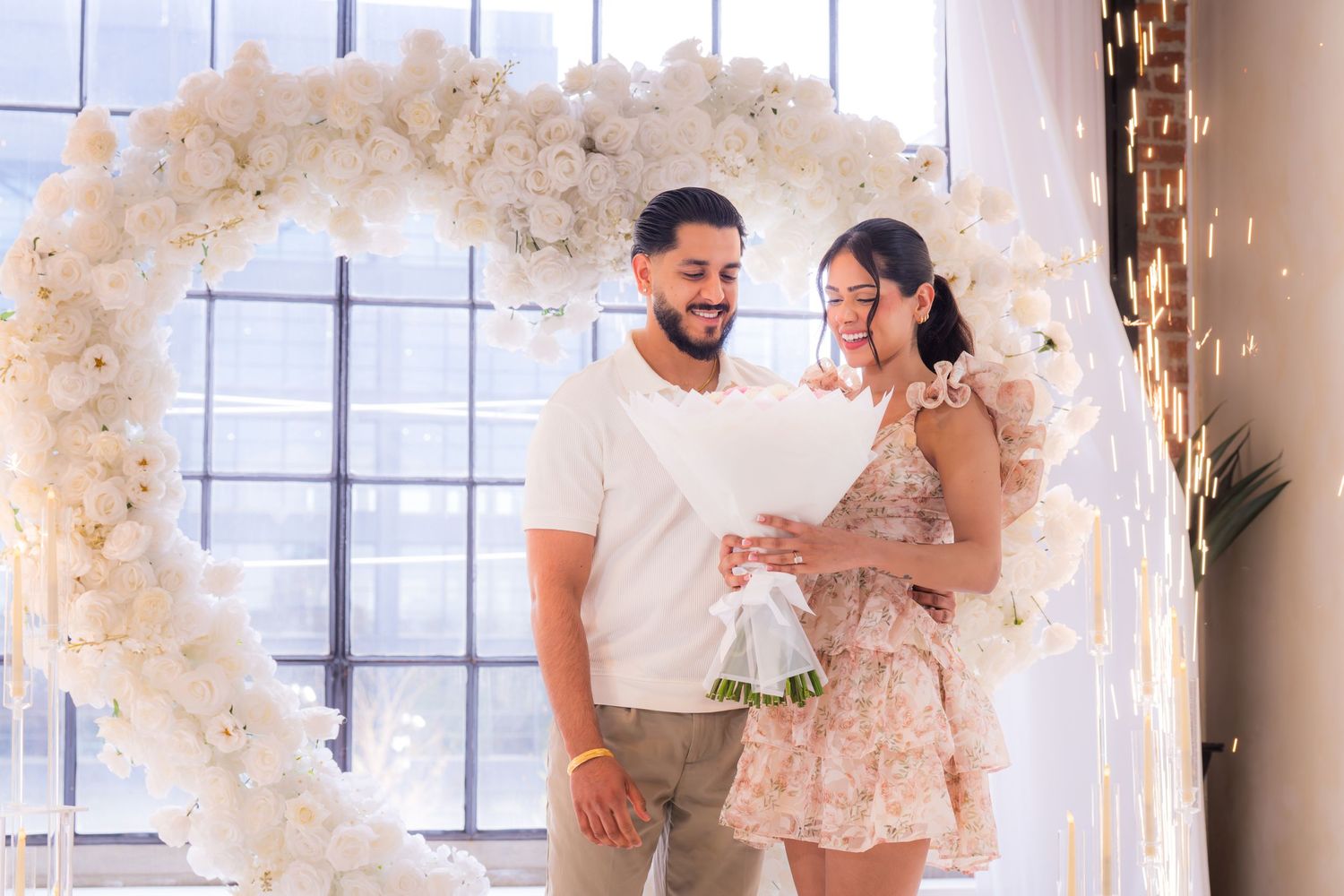 Happy couple standing together under white floral heart arch holding bouquet after proposal.