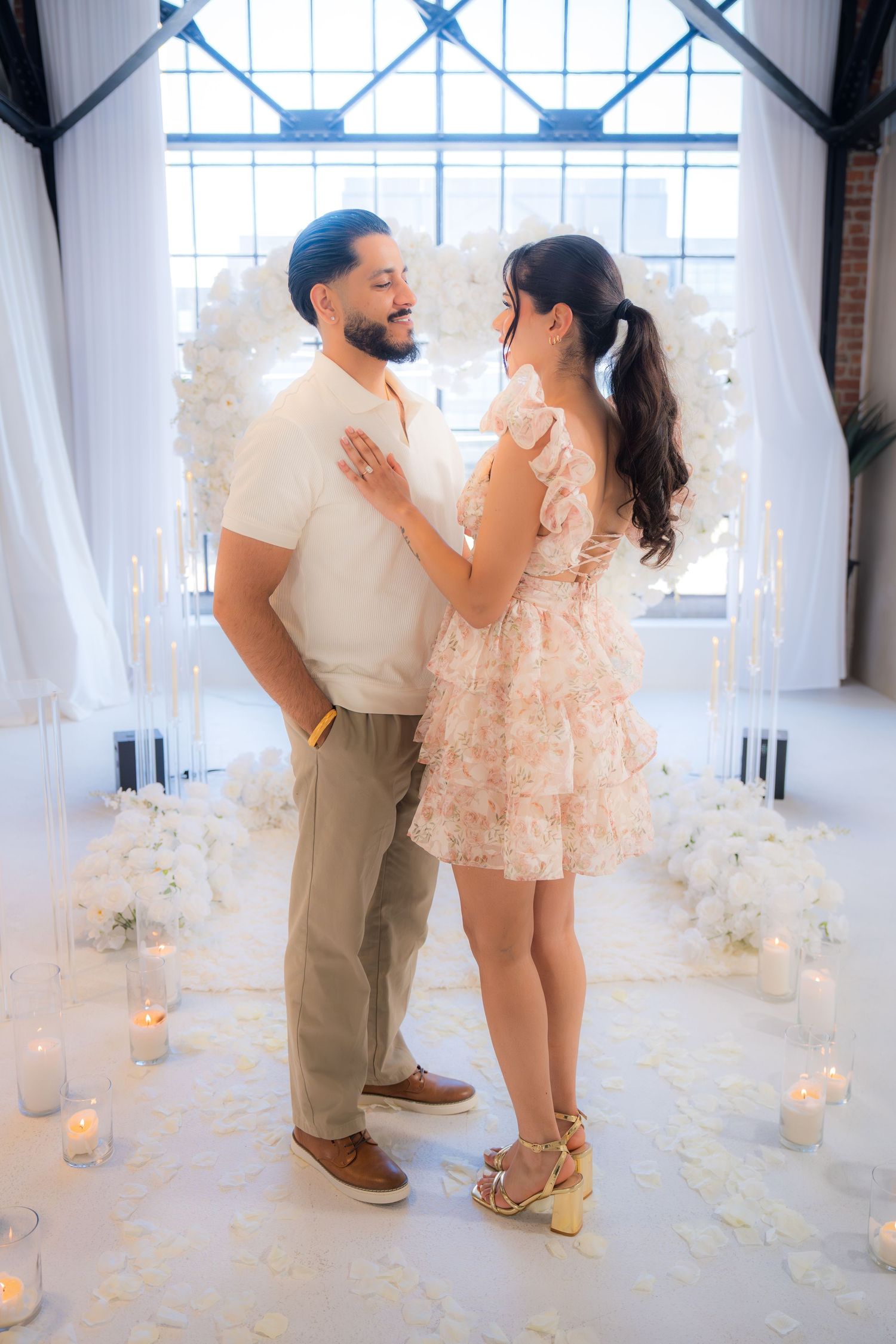 Couple standing face to face in romantic venue with candles and white floral heart arch backdrop.