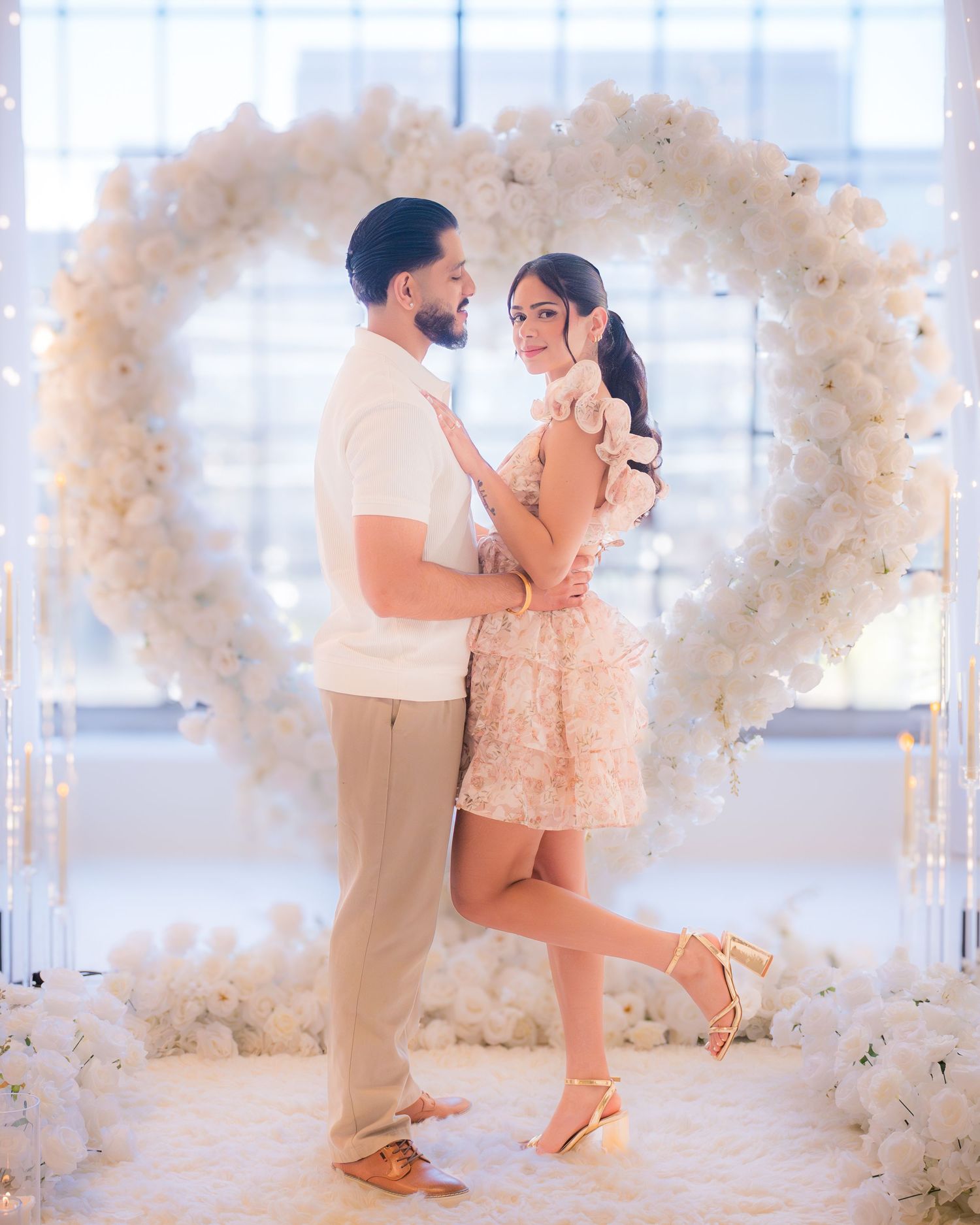 Elegant couple posing in front of a large white floral heart arch in a bright venue.