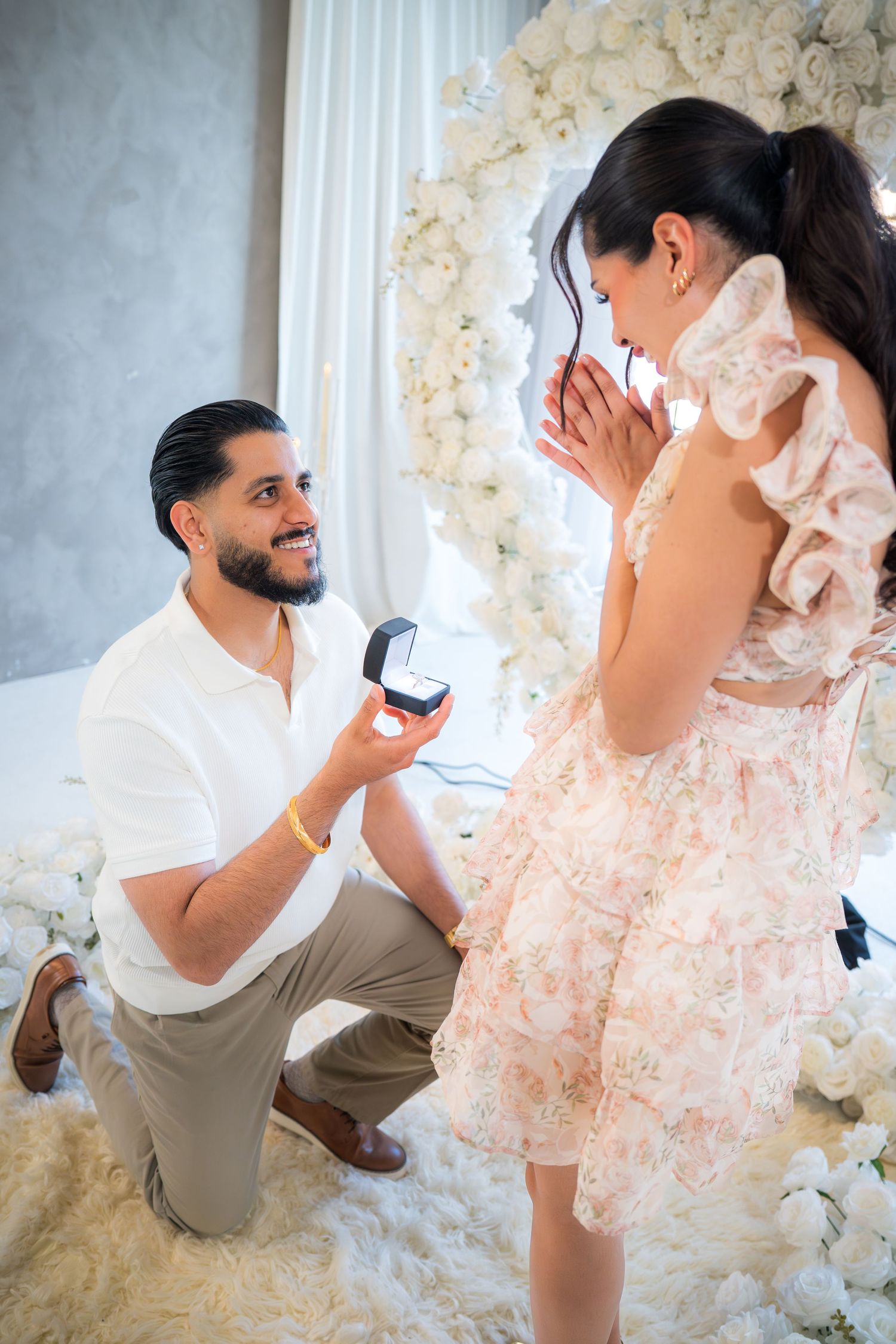 Man proposing on one knee with ring box to woman in floral dress amid white flowers.