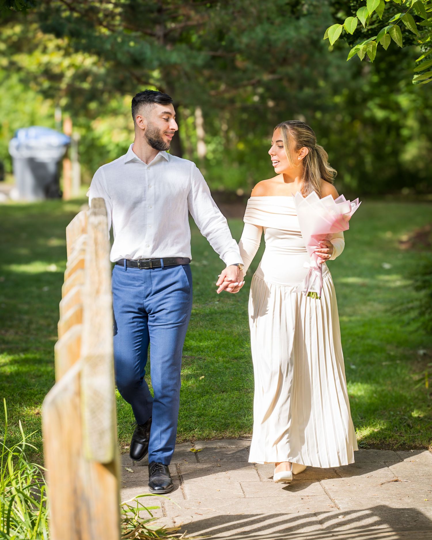 A couple strolling along a garden path with sunlit trees in the background.