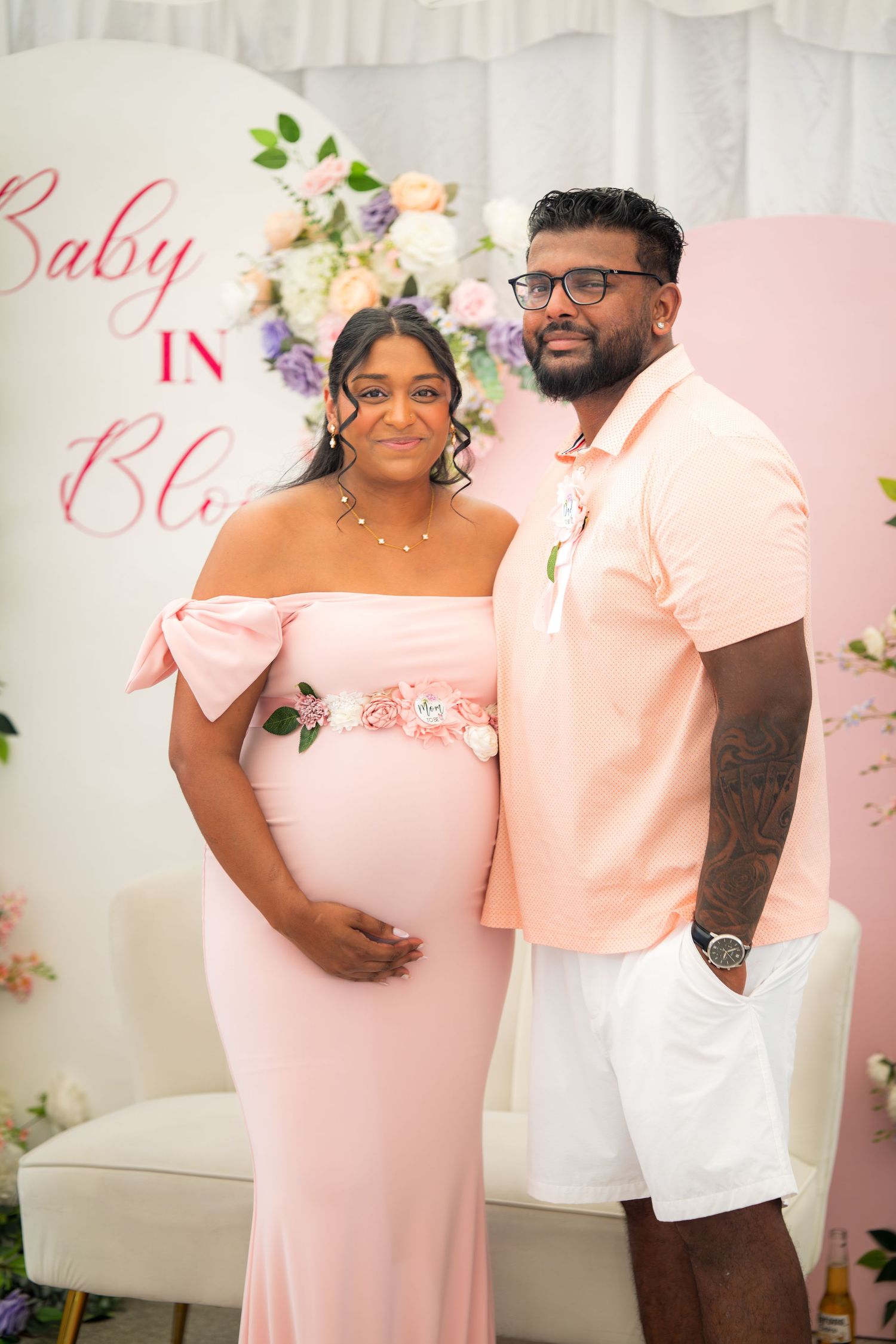 Couple stands together at baby celebration with pink floral backdrop and matching outfits.
