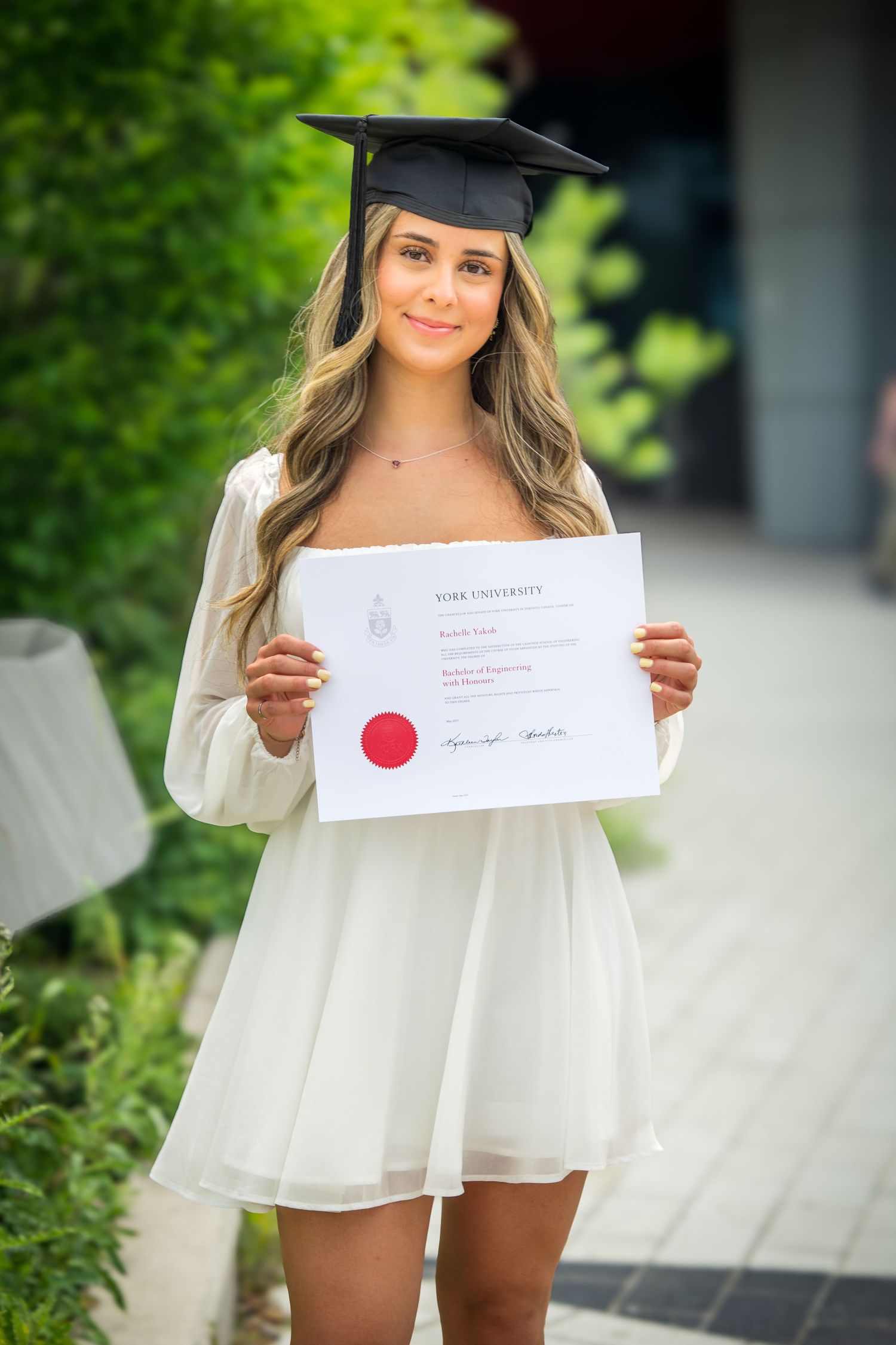 Graduate in white dress displays diploma with red seal outside modern building.