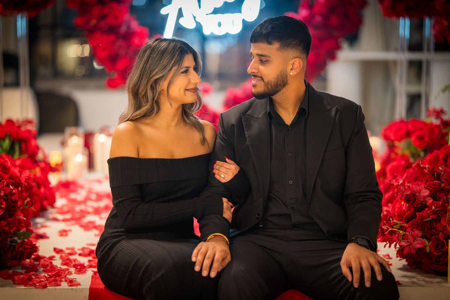 Couple shares joyful moment while sitting against backdrop of red roses and candles.