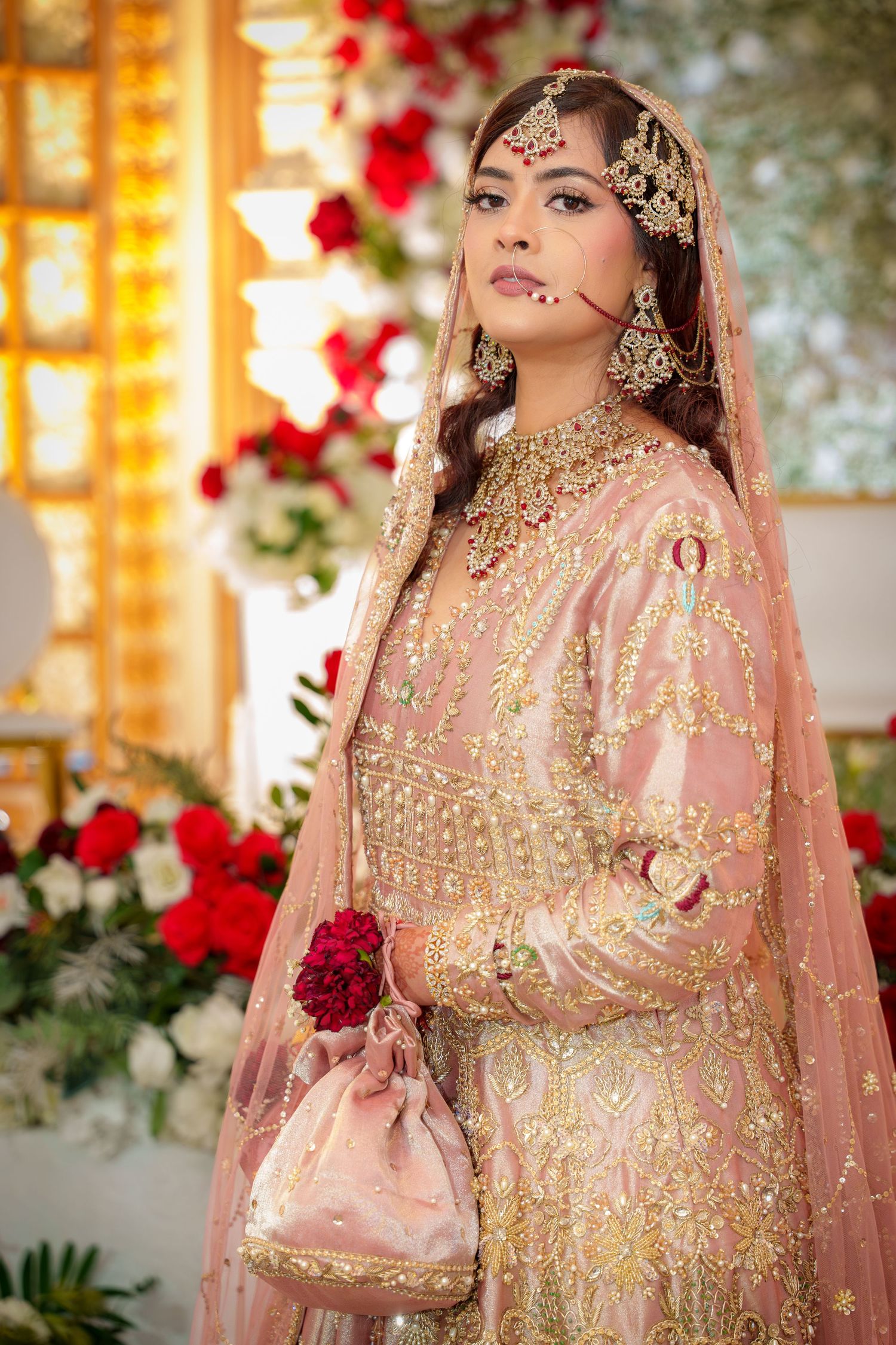 Traditional South Asian bridal attire in peach and gold with intricate embroidery and jewelry against floral decor.
