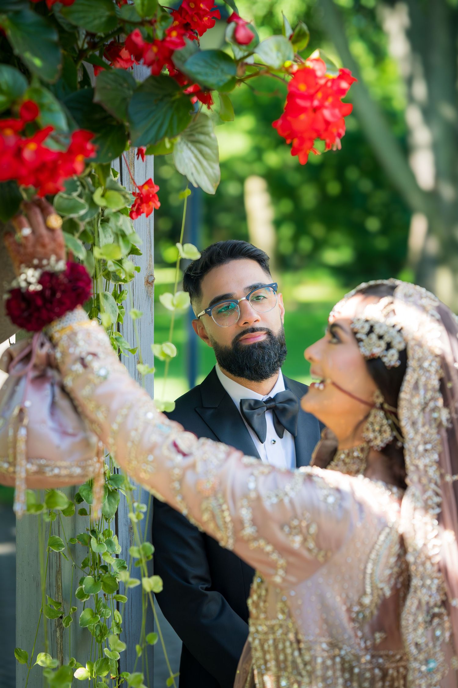 A couple in traditional wedding attire poses under red flowers in a garden setting.