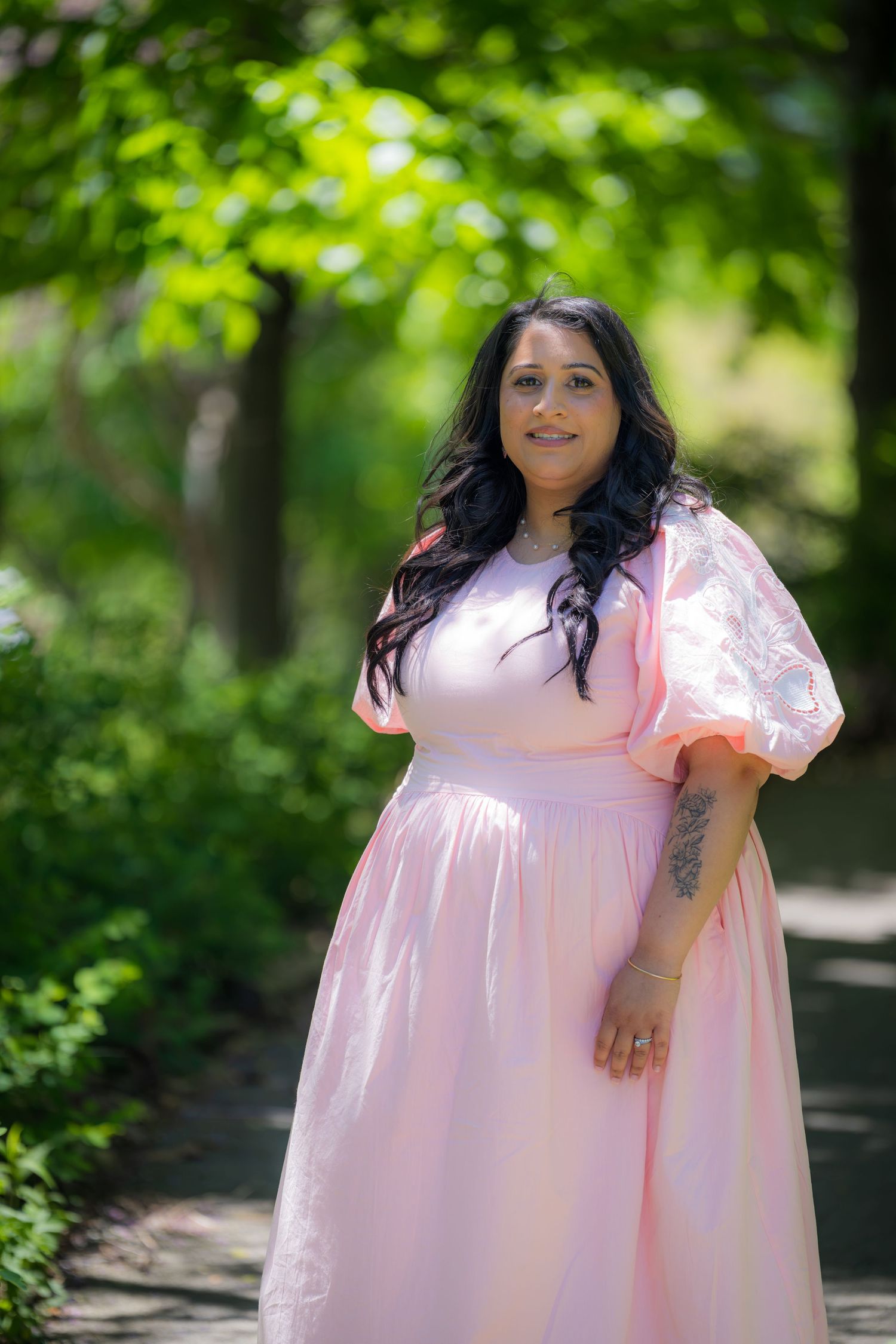 Woman in elegant pink puff sleeve maxi dress poses on garden path surrounded by greenery.