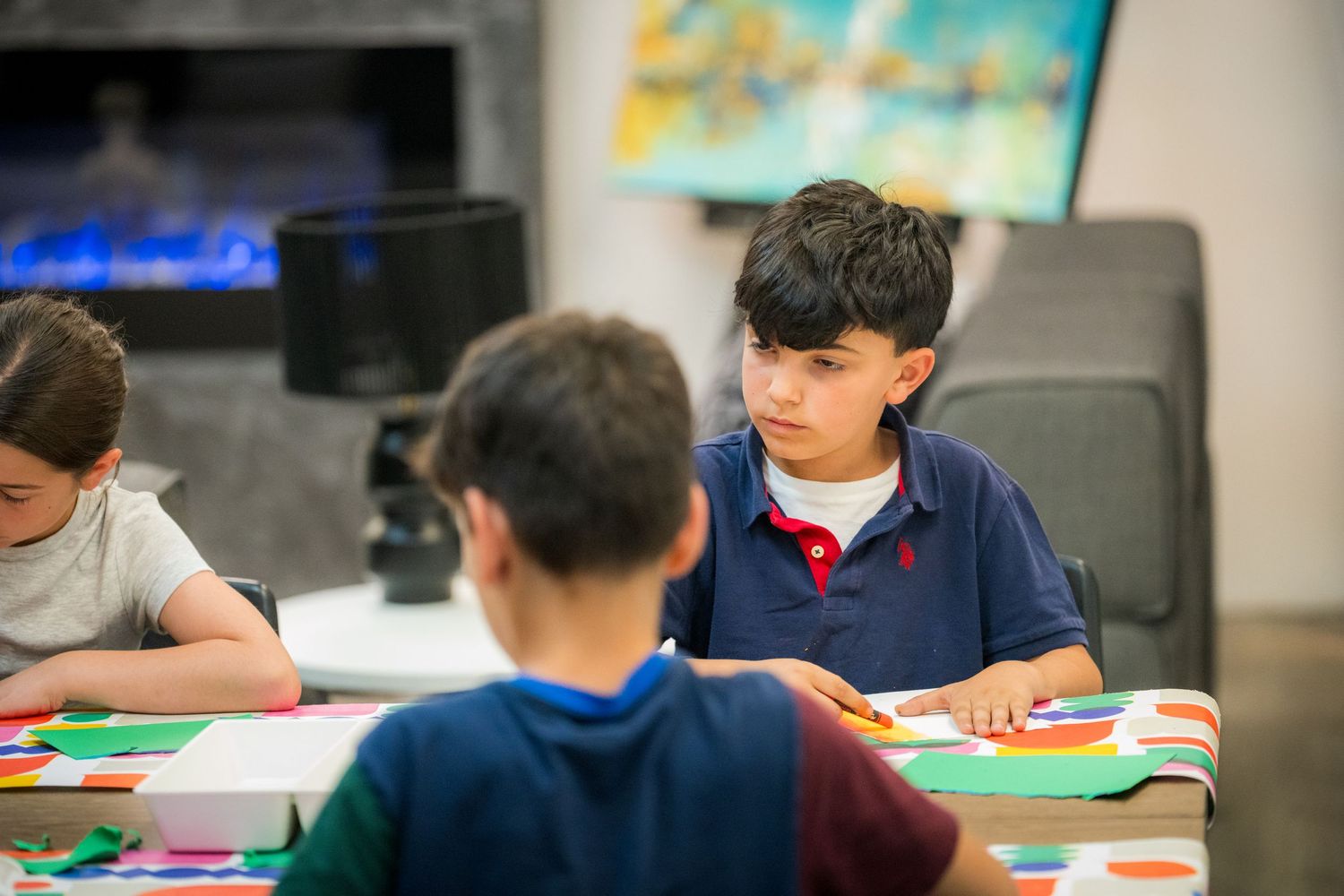 Young boy in blue polo shirt looks up during colorful paper craft activity at art workshop table.