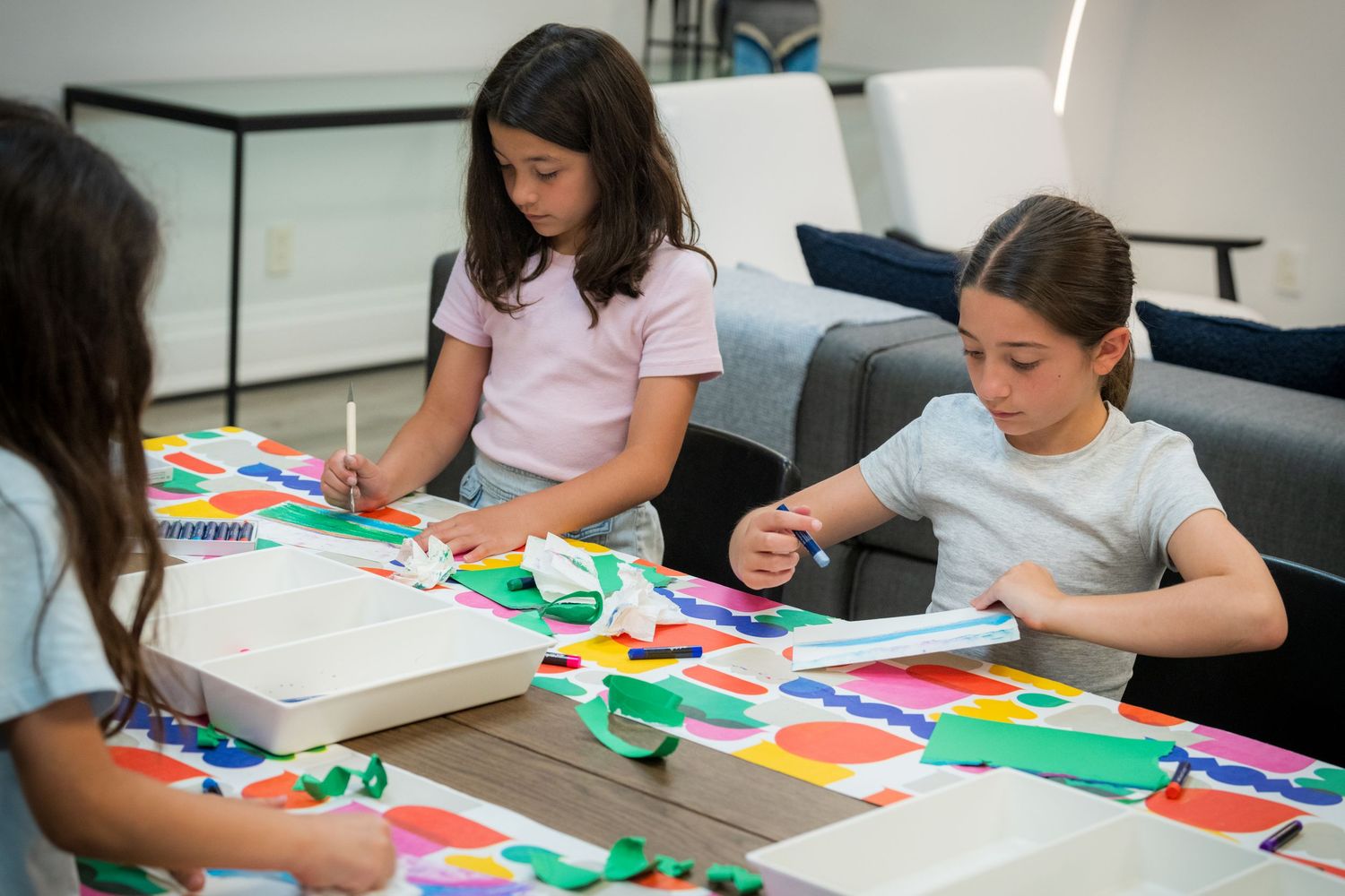 Three girls collaborate on colorful paper collage art projects at a shared table.