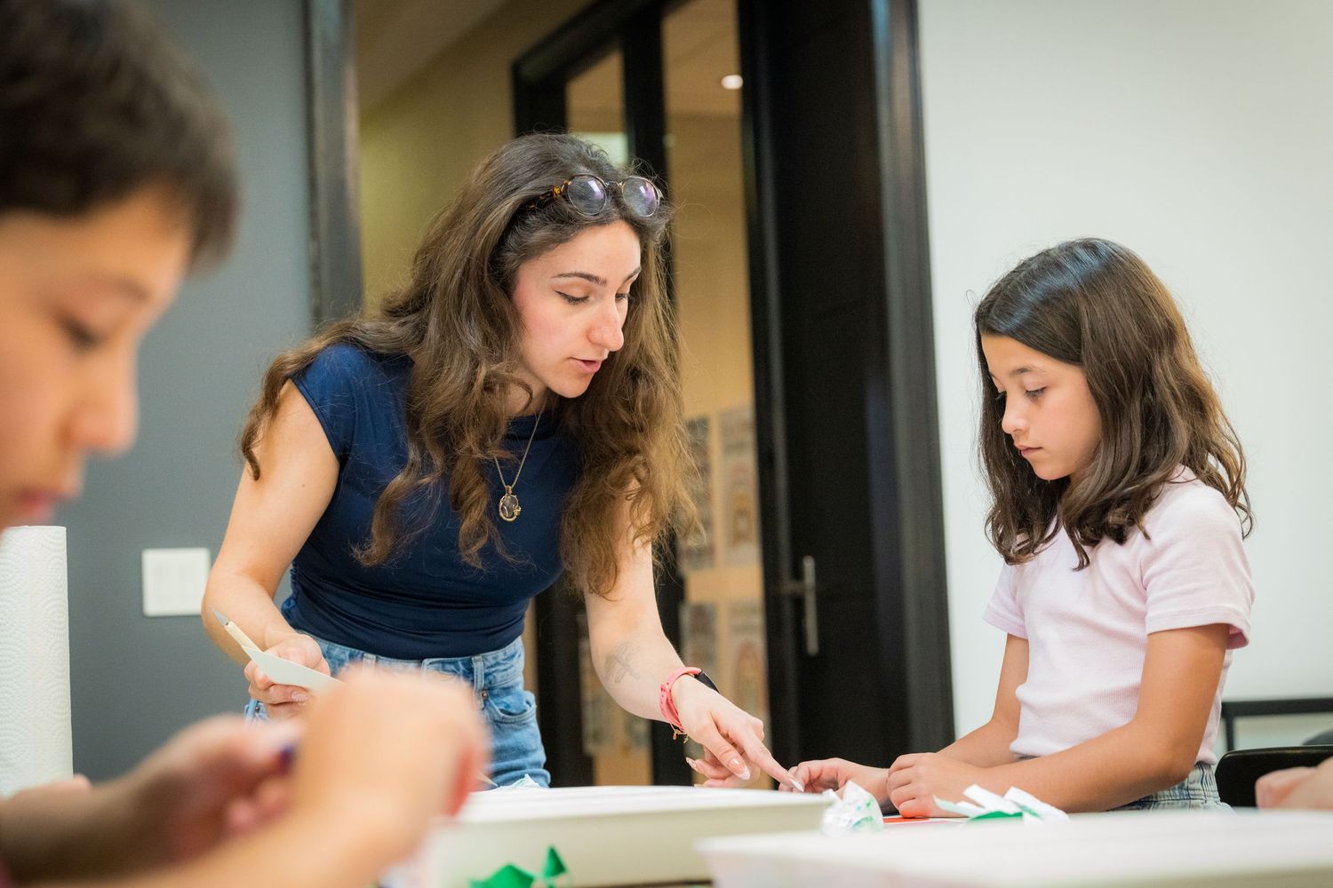 A female art instructor helps a young girl with a hands-on paper craft project at a table.