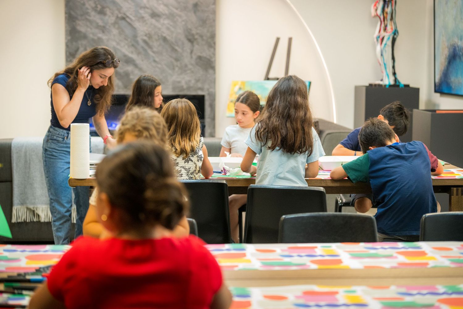 Children engaged in an art activity at tables in a bright classroom, supervised by a young female teacher.
