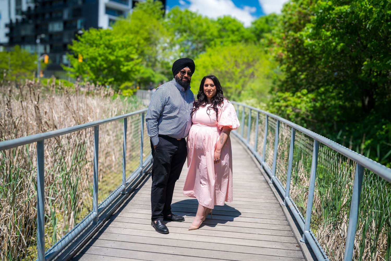 Couple poses on a wooden bridge surrounded by lush greenery, woman in pink maternity dress, man in gray suit.