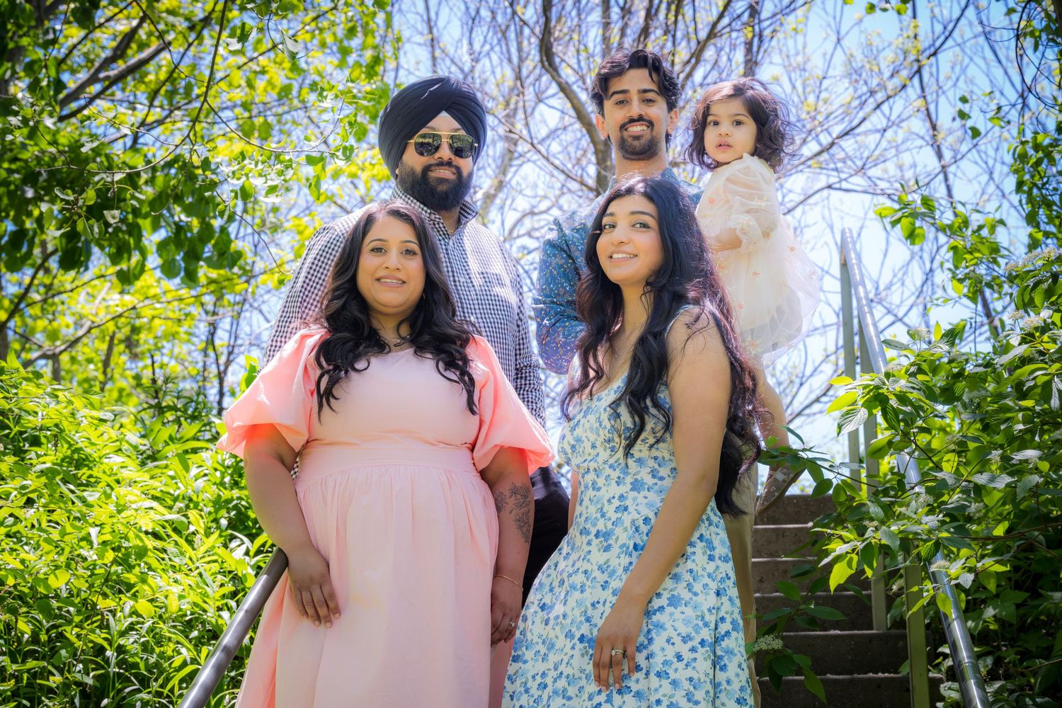 Extended Sikh family group posing on outdoor steps surrounded by lush green trees in sunlight.