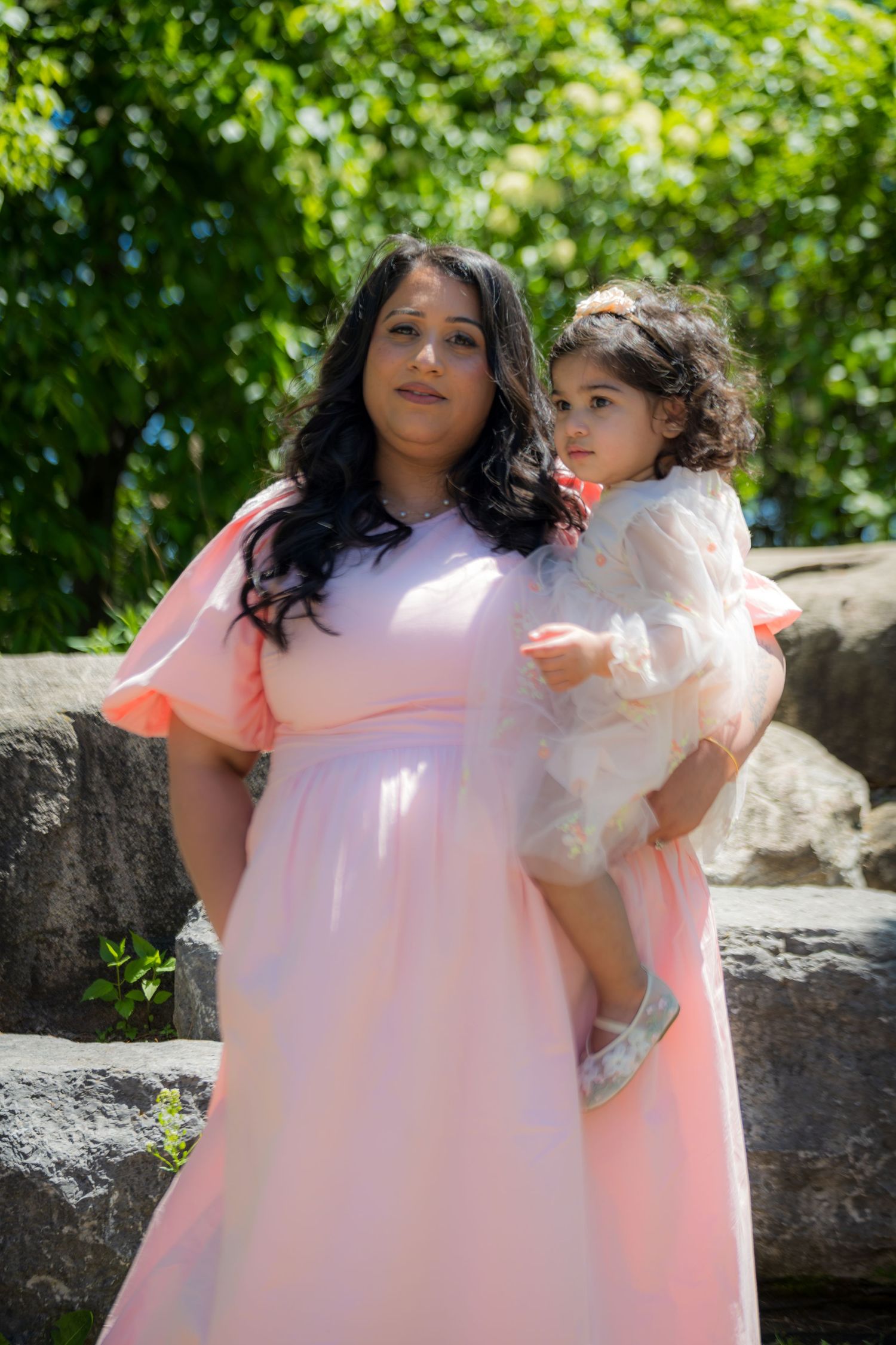 A woman in a pink maxi dress holds a young toddler girl in a white dress outdoors near stone steps.